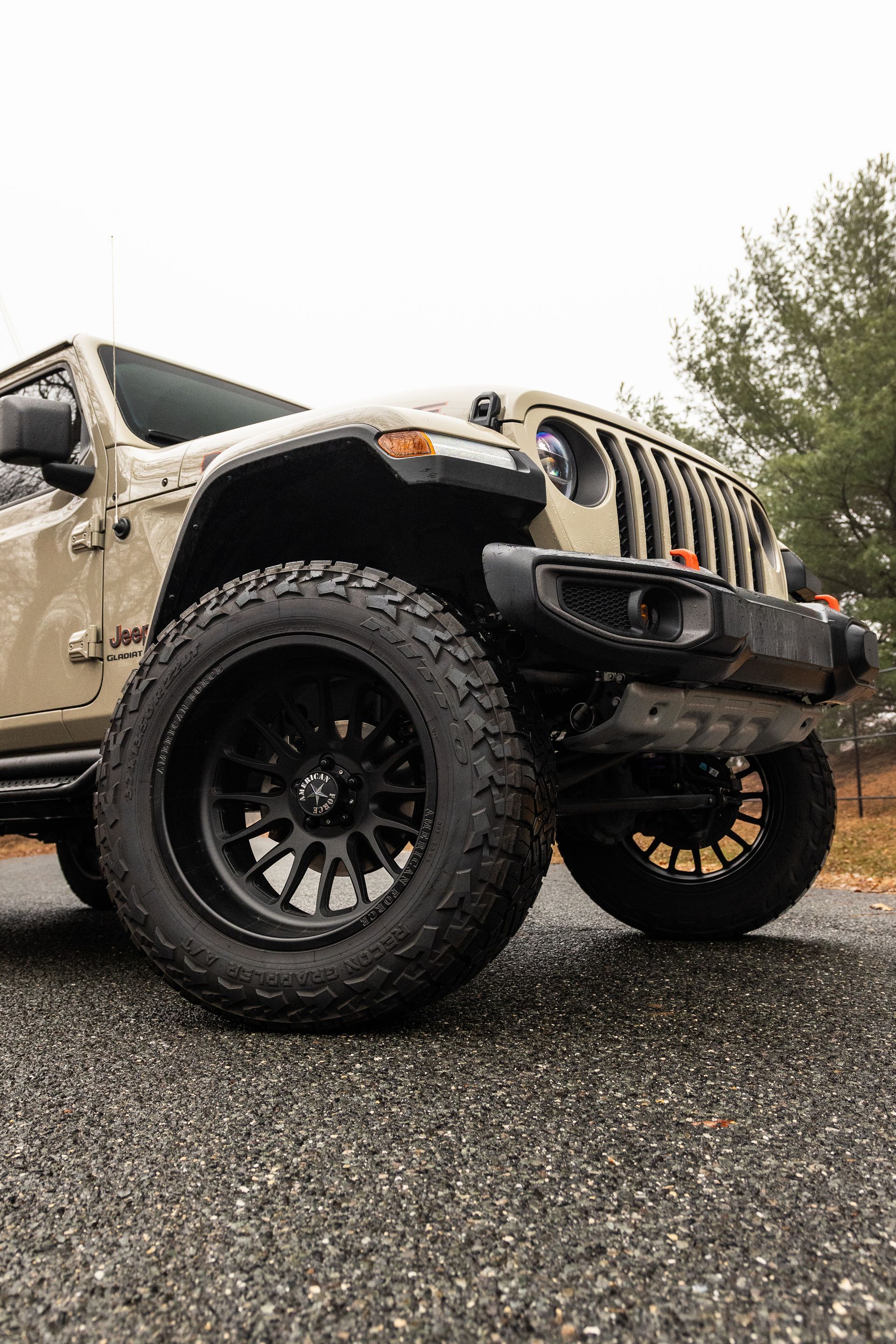 Tan Jeep Wrangler with black wheels, parked on asphalt, cloudy sky.