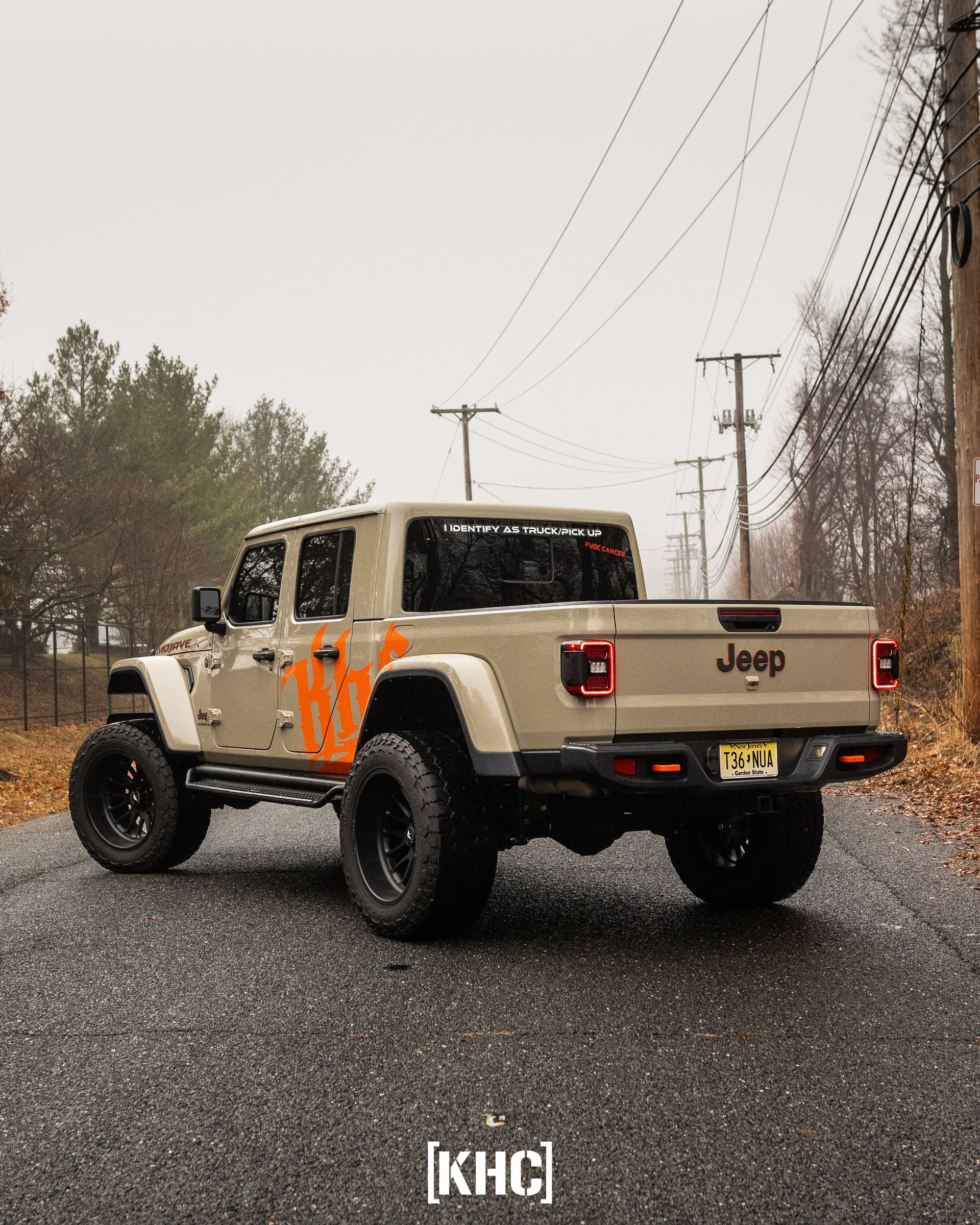 Tan Jeep Gladiator truck on a wet road, with large black wheels and orange accents on a cloudy day.