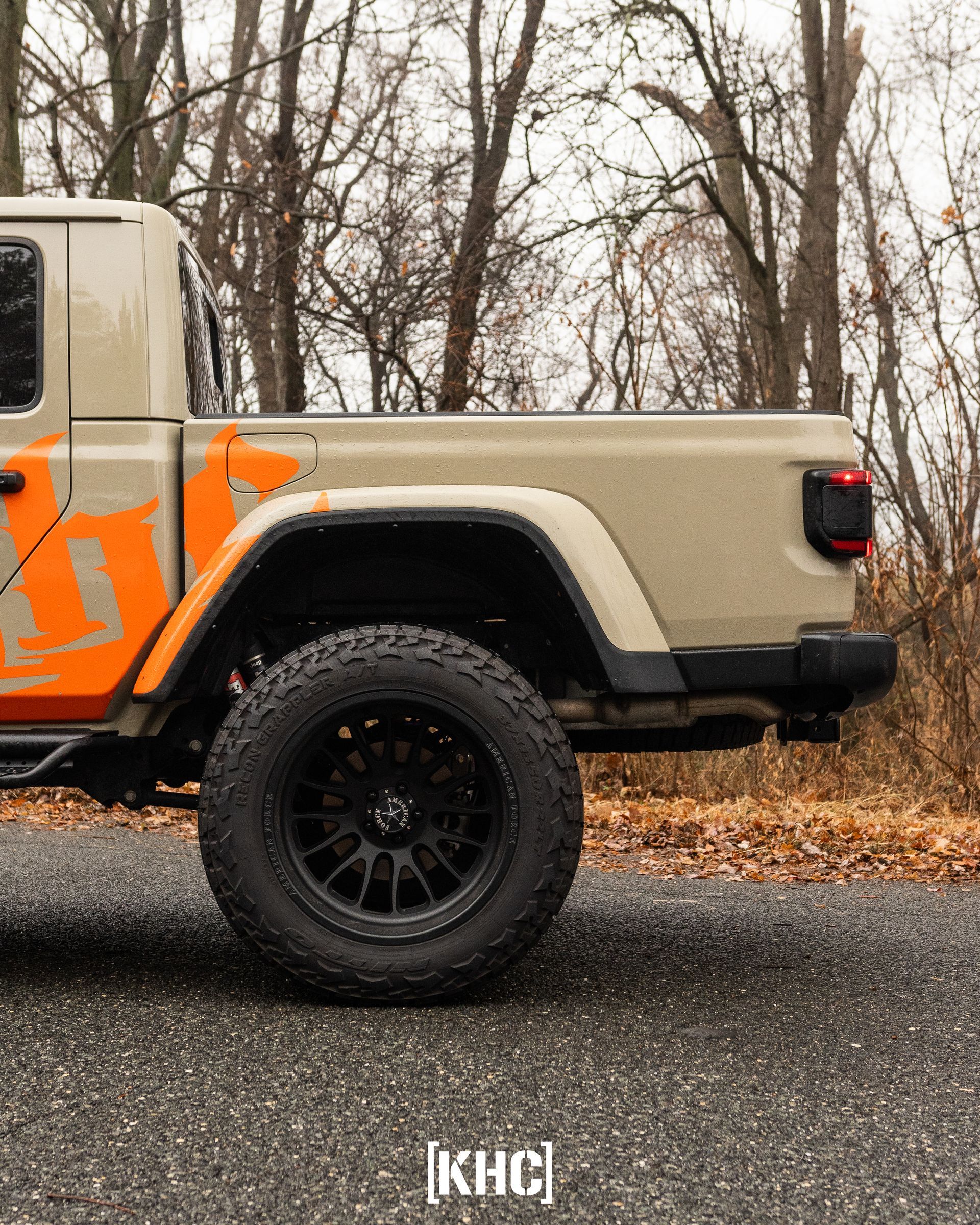 Tan Jeep truck with black wheels parked on a road, orange logo on the side, forest background.