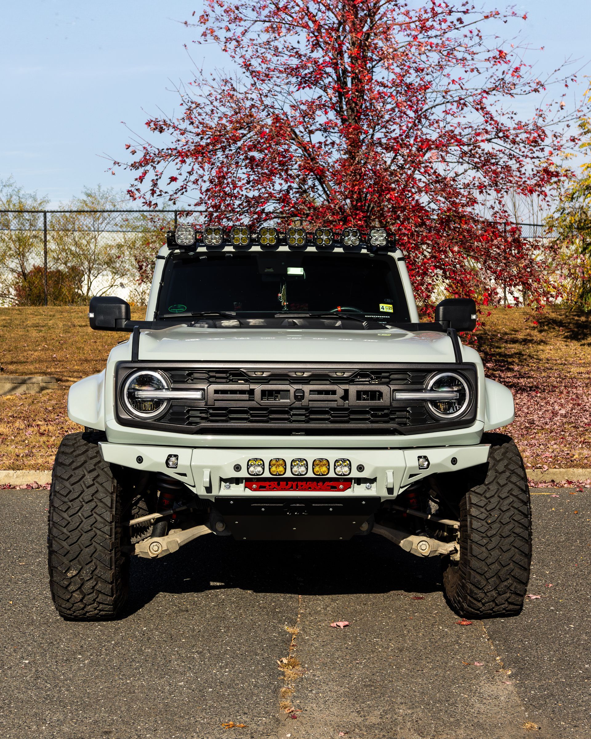Gray Ford Bronco with black accessories, large tires, and LED lights, parked in front of red-leafed tree.