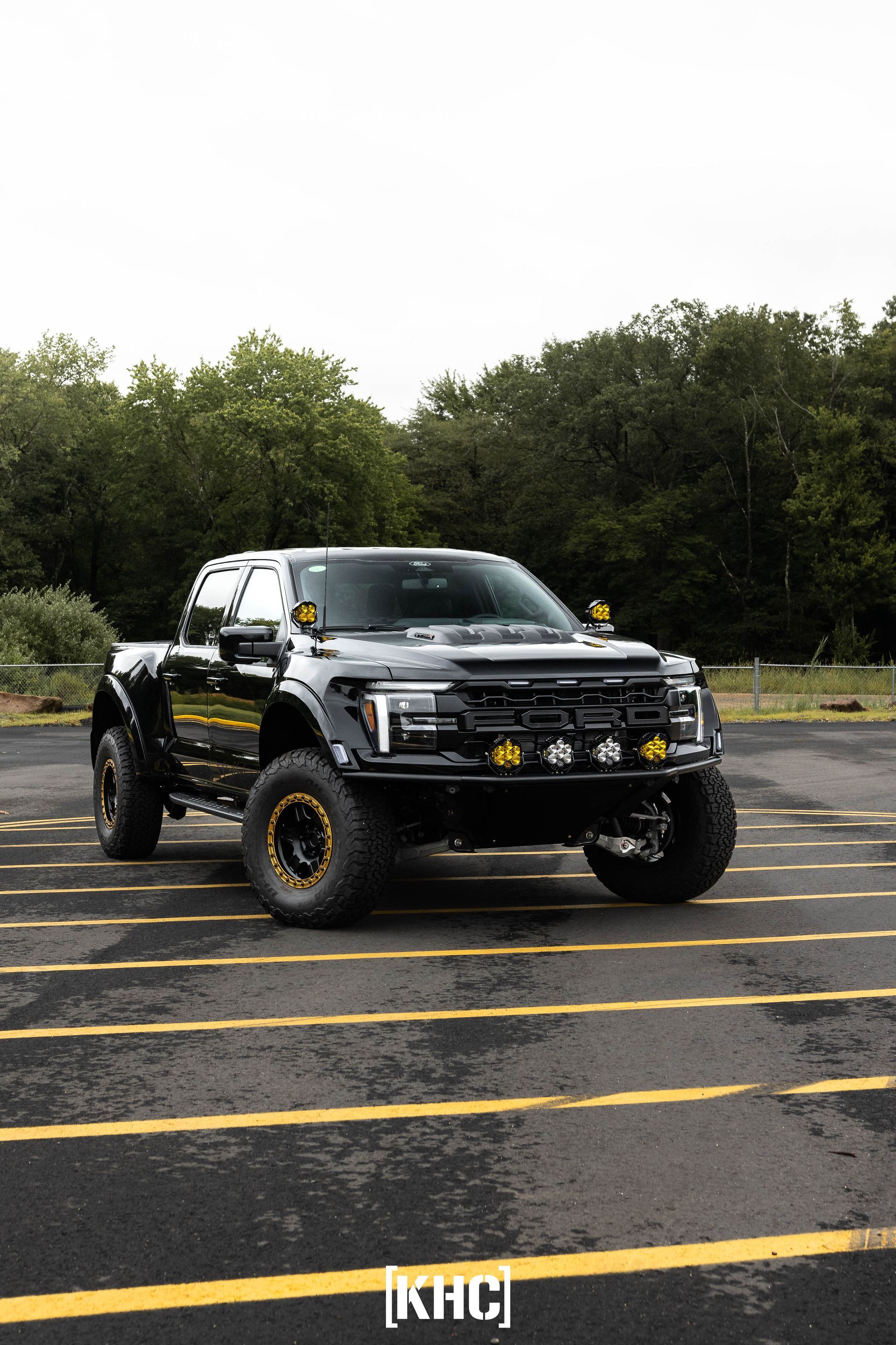 Black lifted Ford F-150 Raptor with yellow accents parked on asphalt in front of trees.
