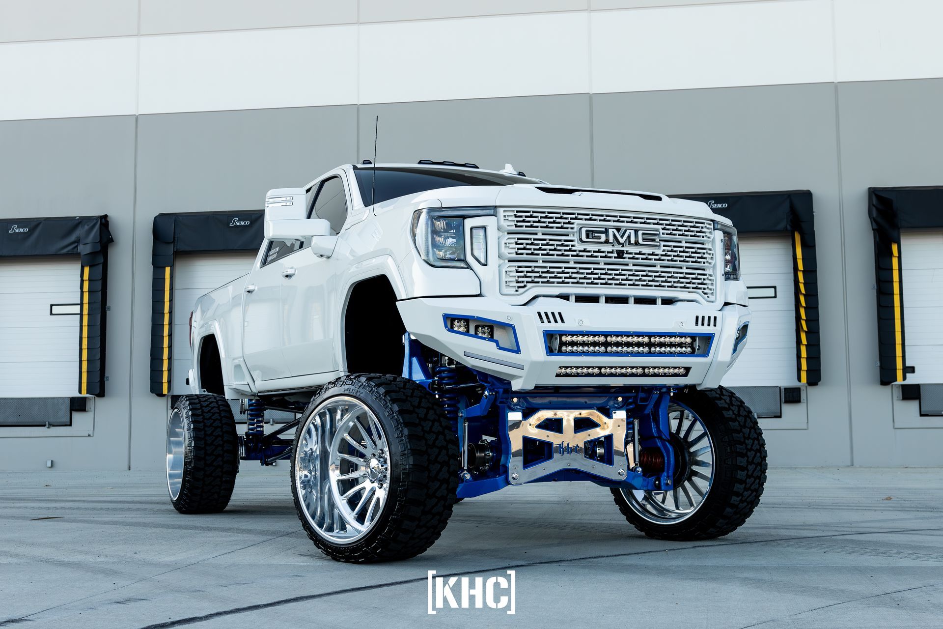 White and blue lifted GMC truck with large chrome wheels parked in front of loading docks.