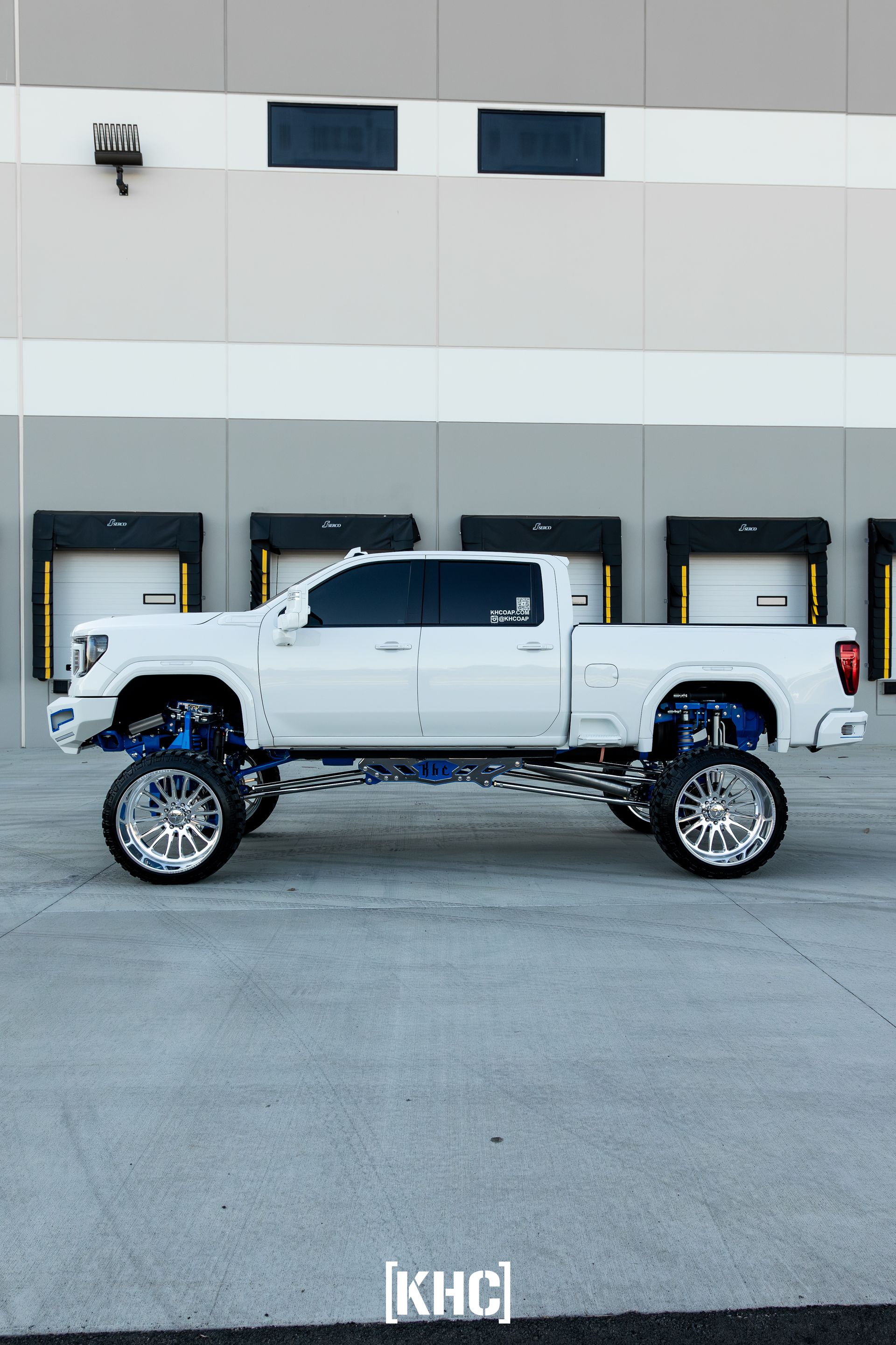 White lifted truck with blue suspension, chrome wheels, parked in front of a warehouse.