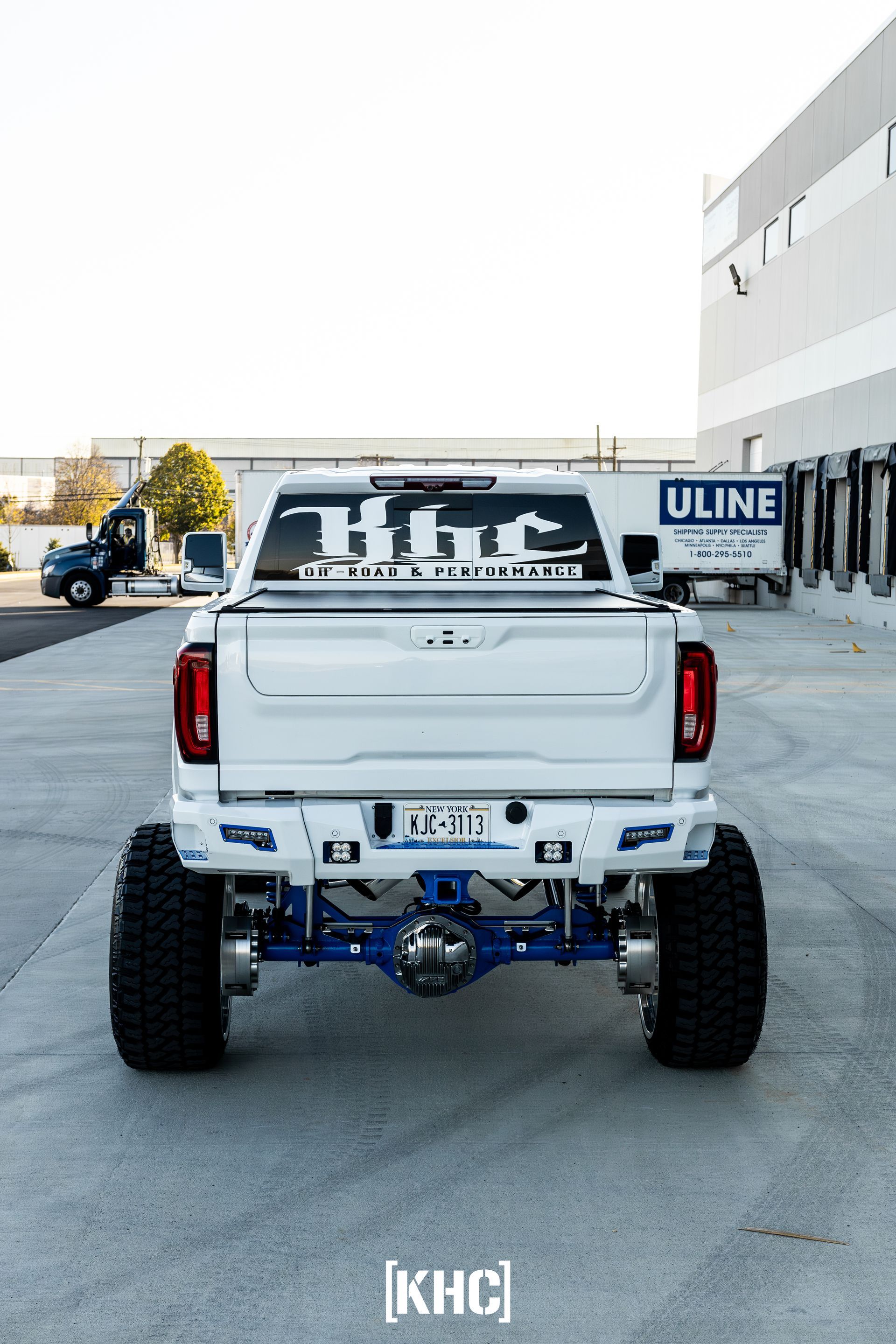 White truck with large tires, blue suspension, parked near a warehouse.