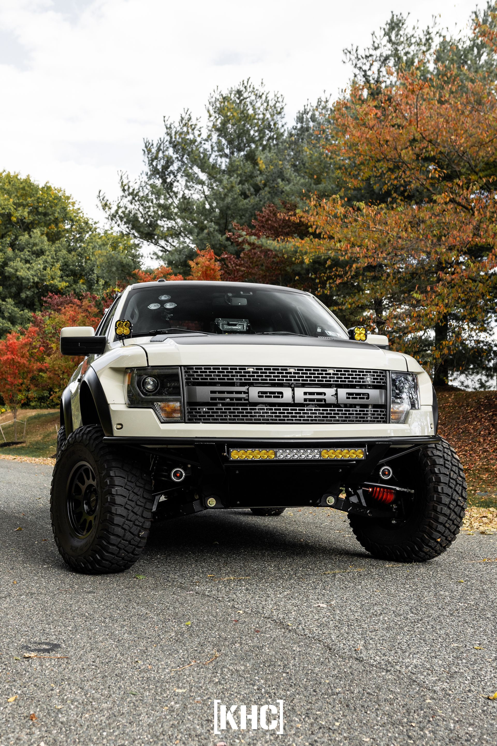 White lifted Ford Raptor truck, black wheels, off-road lighting, parked in front of autumn trees.