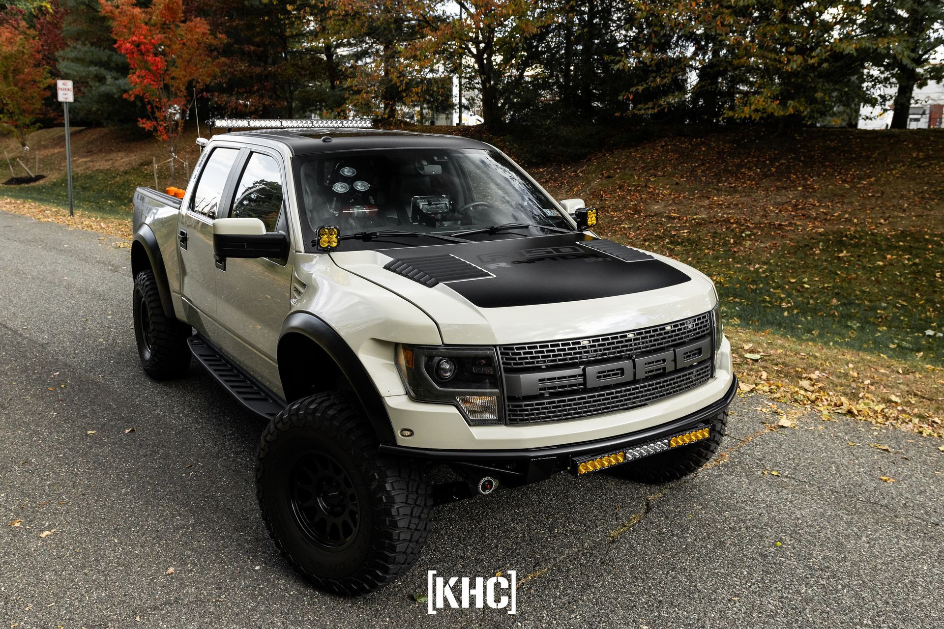 White Ford Raptor truck with black accents parked on a road with fall foliage.
