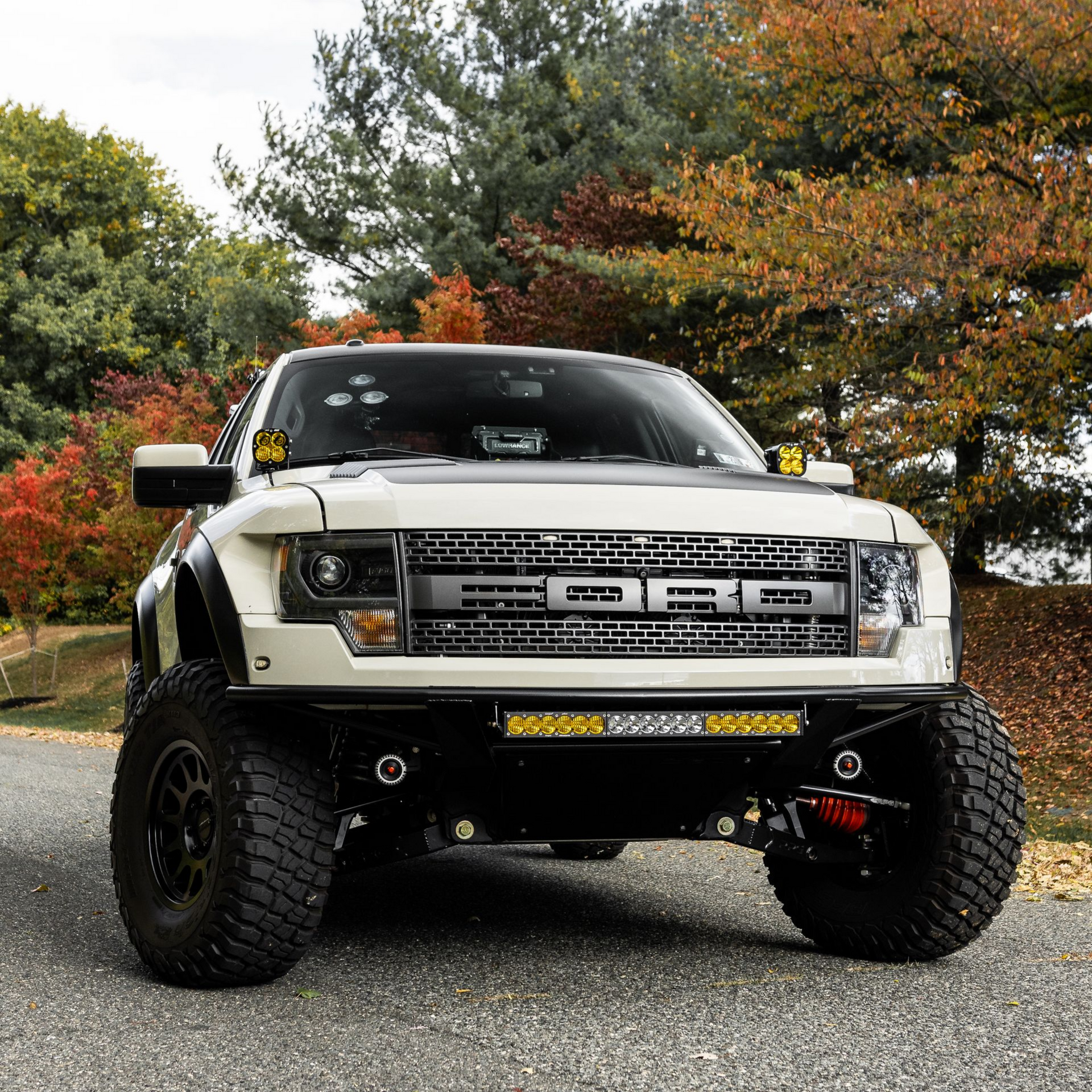 White Ford Raptor truck with black accents, large tires, and off-road lights parked on a road with fall foliage in the background.