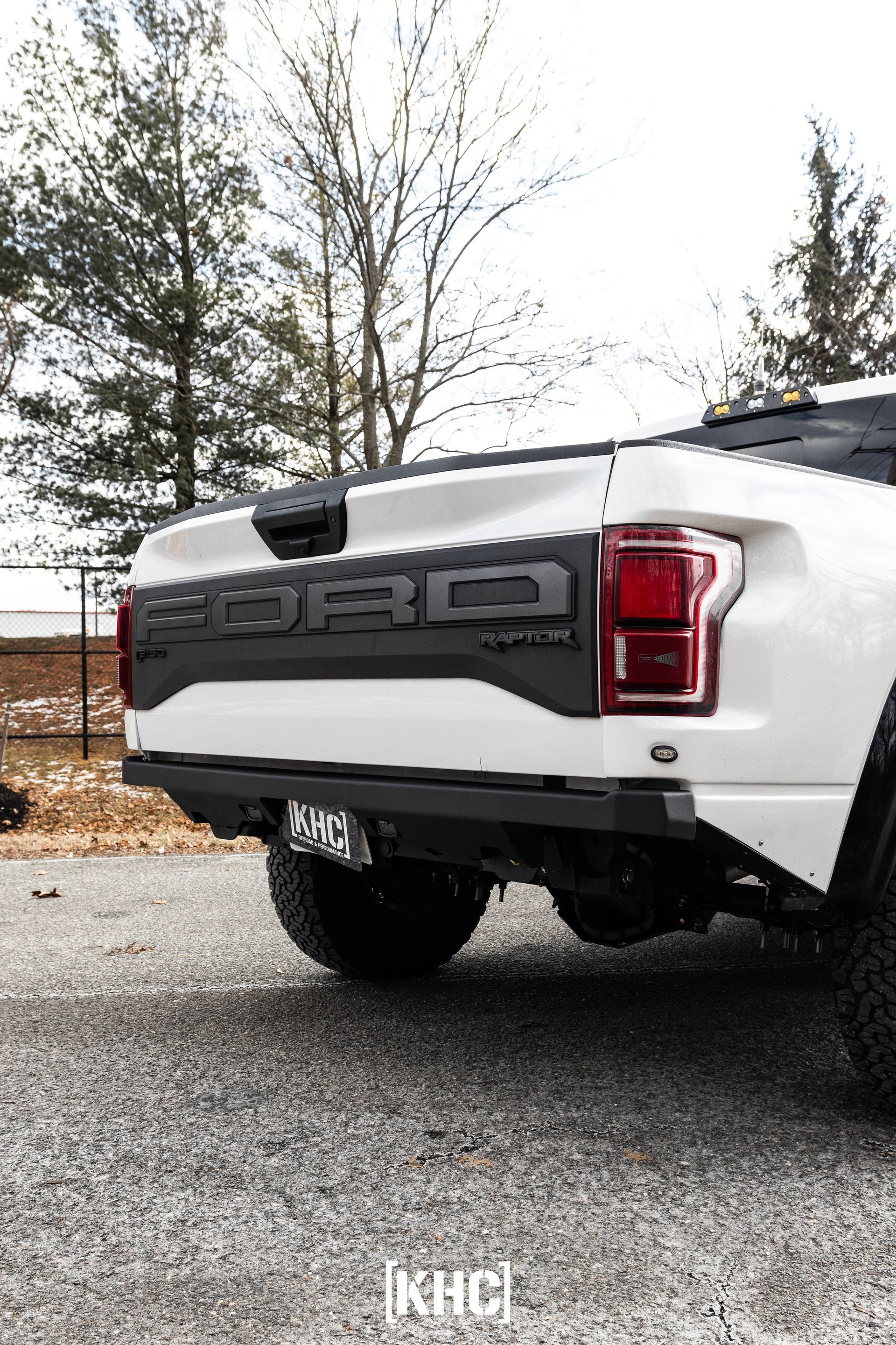 White Ford Raptor truck's rear with black accents; parked outside, trees in background.
