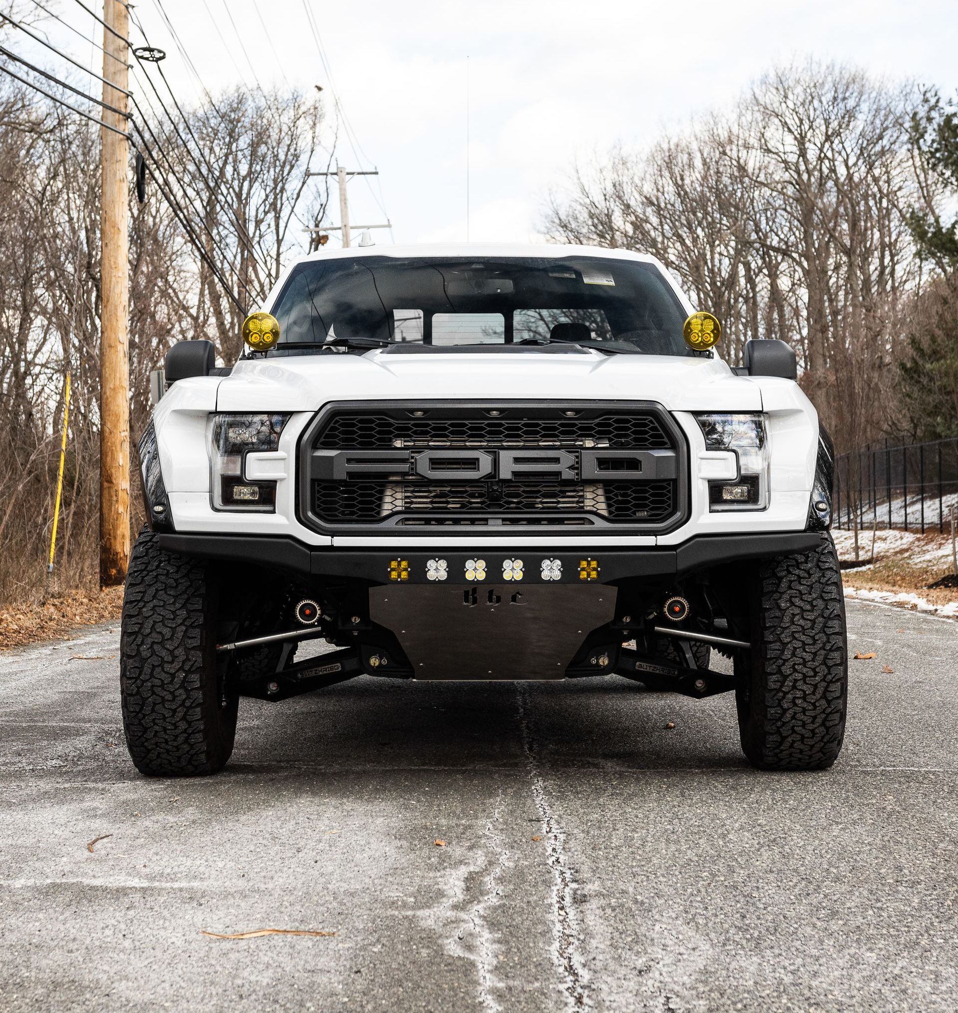 White Ford Raptor truck on a road, with custom front bumper, off-road lights, and black accents.