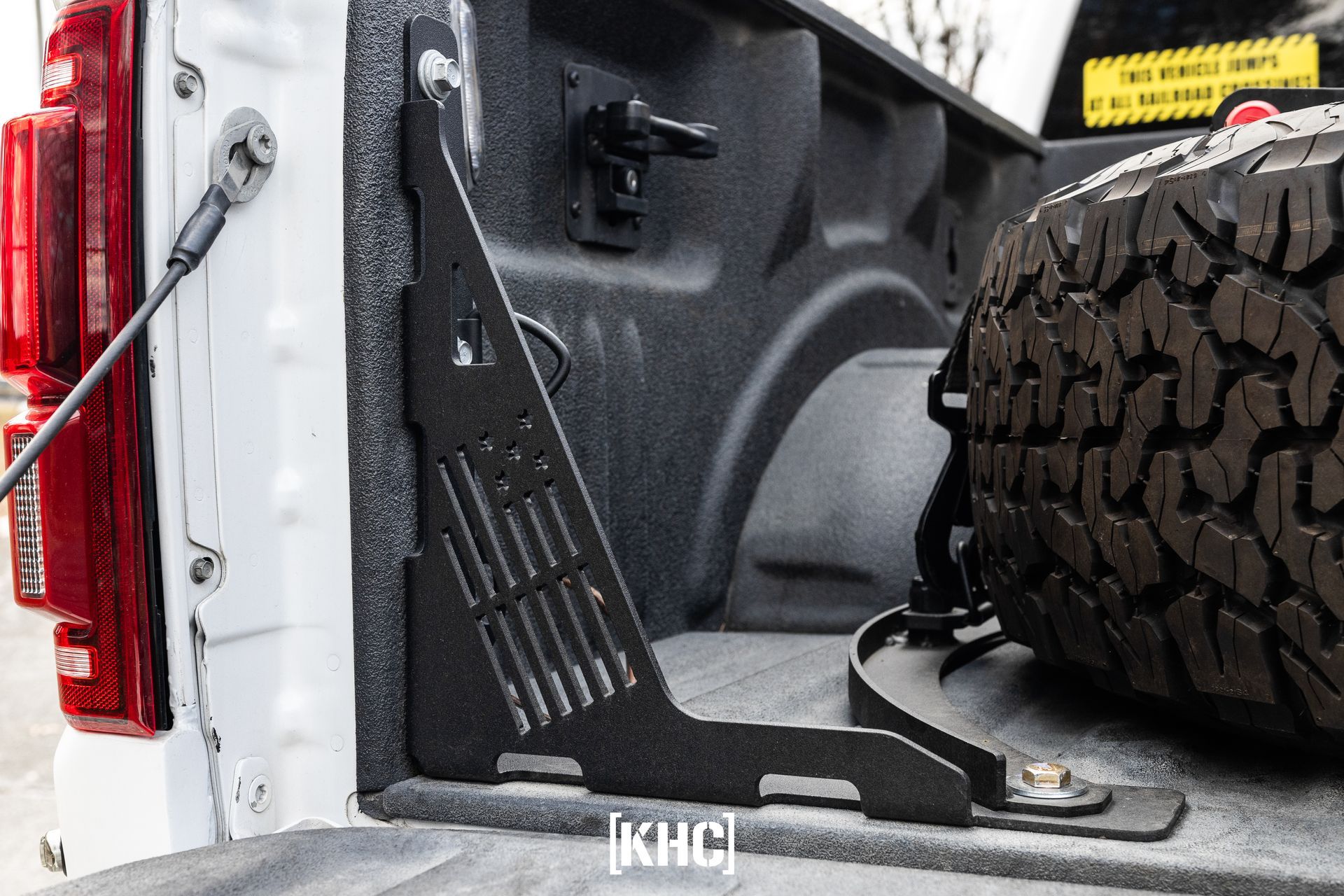 Black metal truck bed mount holding a spare tire, mounted to the truck's sidewall.