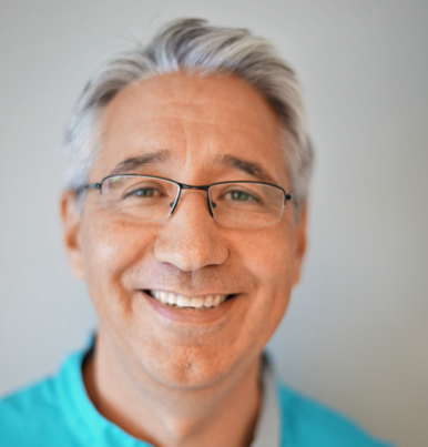 Smilende tannlege i blå skrubber, ved et vindu, med tannverktøy i bakgrunnen.