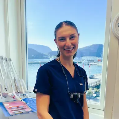 Smilende tannlege i blå skrubber, ved et vindu, med tannverktøy i bakgrunnen.