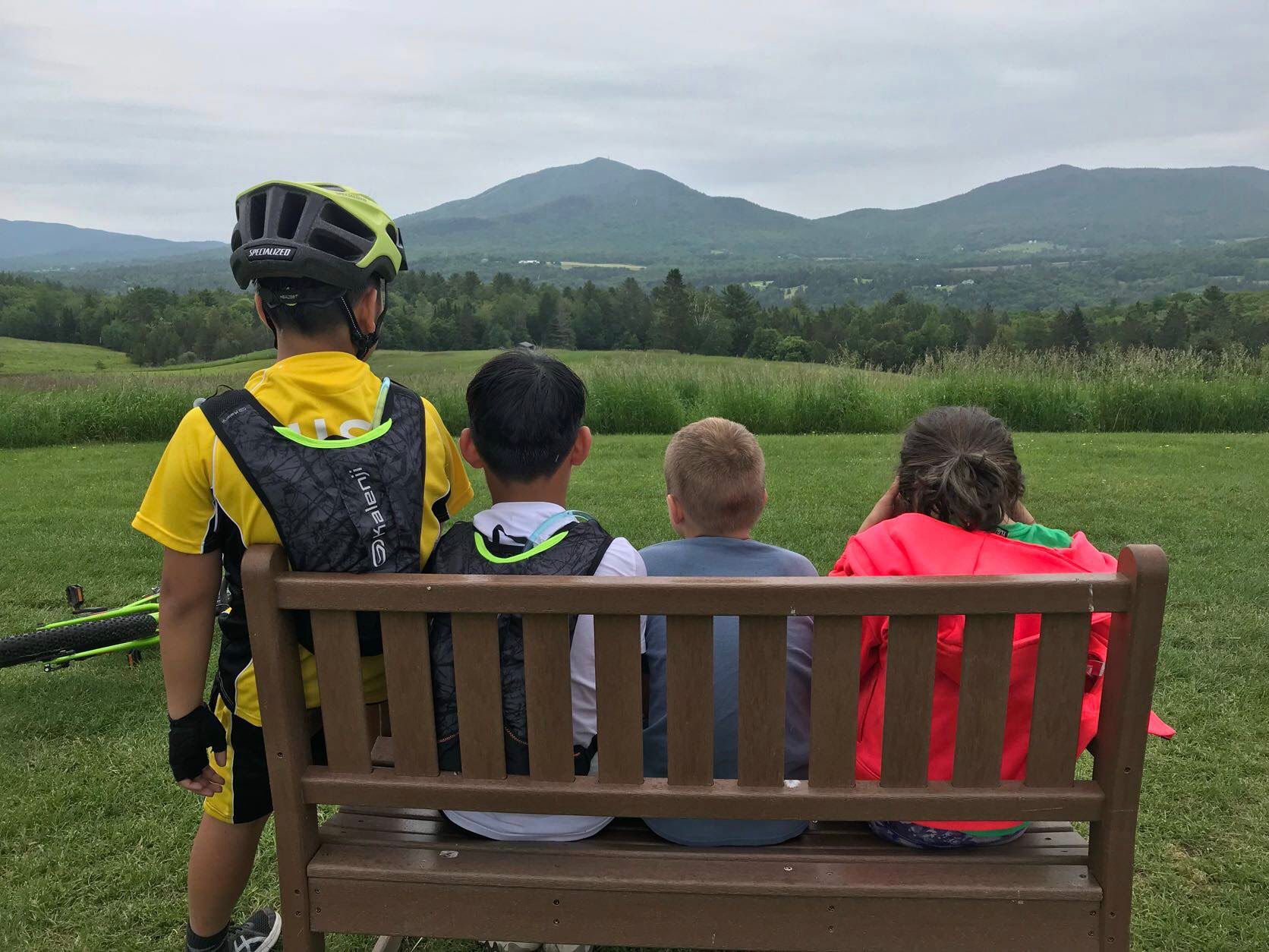 Four kids in bike gear sitting on a bench together in front of a mountain view