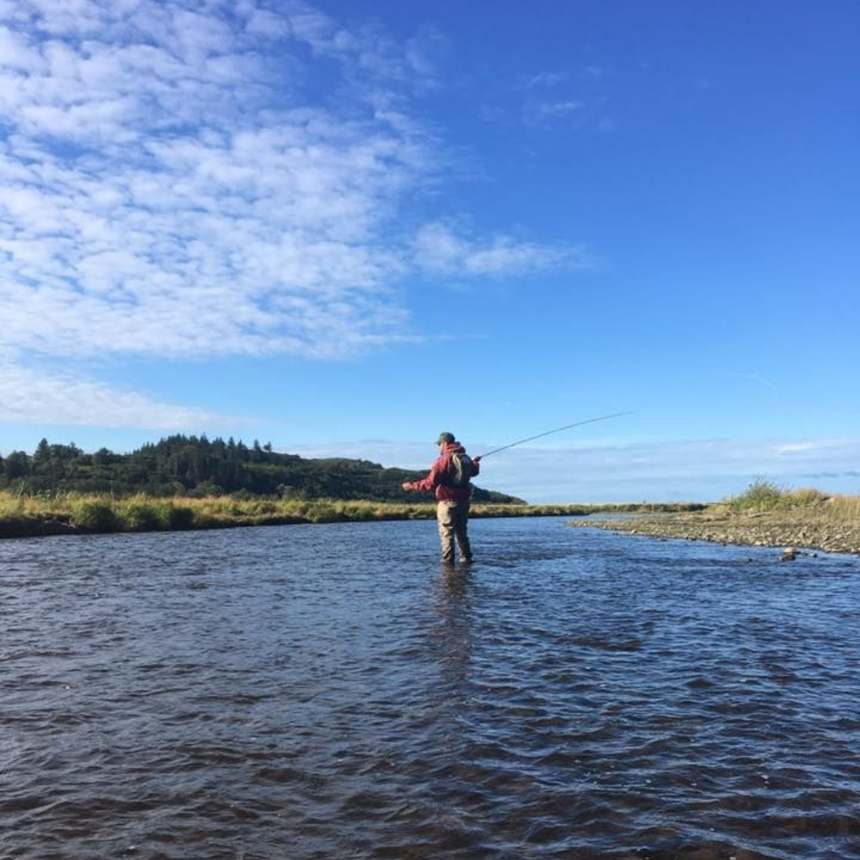 A person in a red jacket fly-fishes while standing mid-stream in a river on a bright, sunny day.