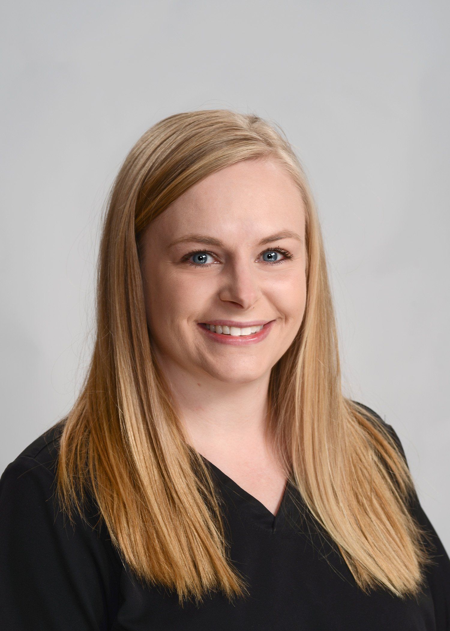 A woman with long blonde hair is wearing a black scrub top and smiling for the camera.