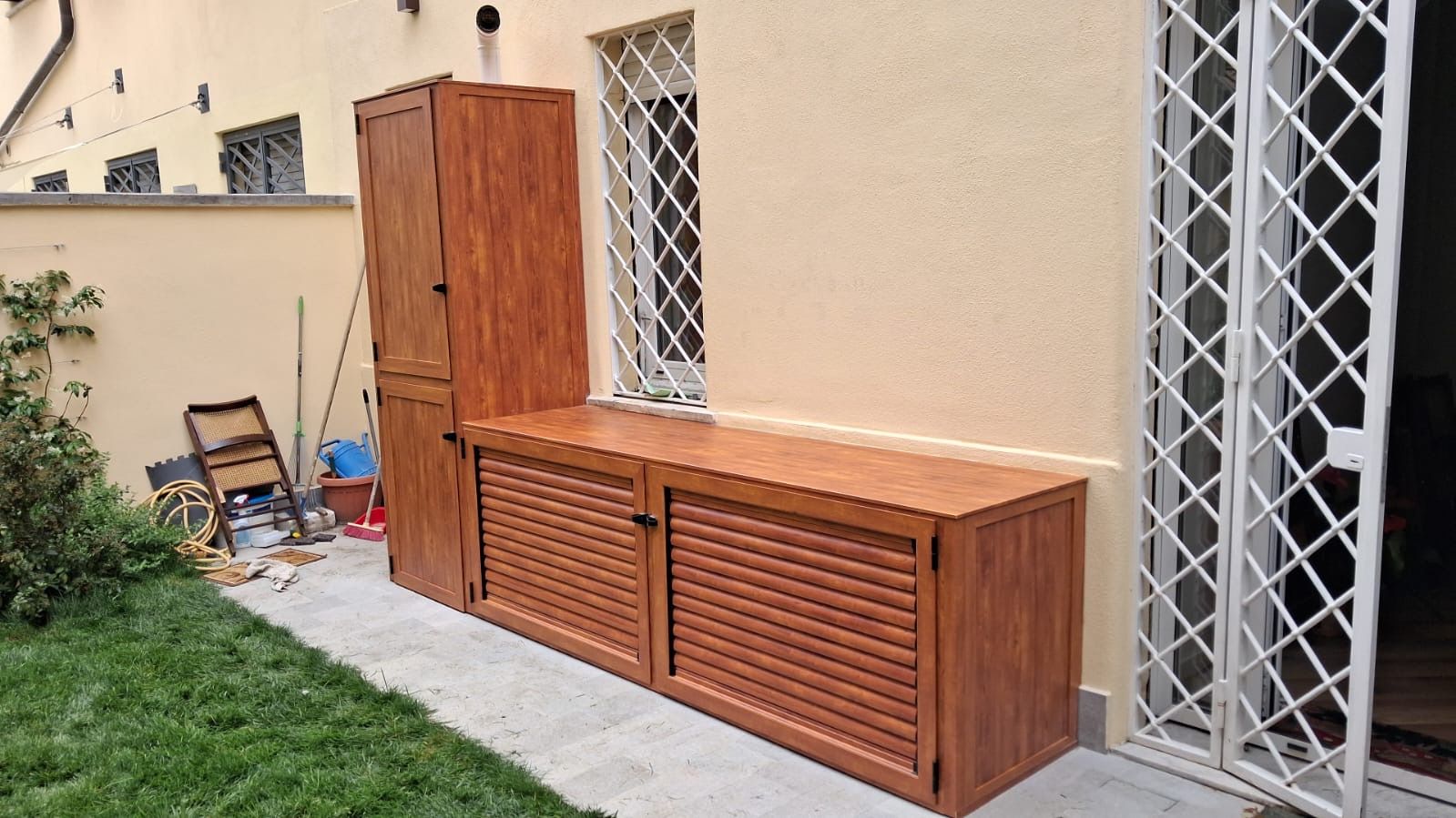 Mobile contenitore da esterno in legno e panca lungo una parete di una casa beige accanto a un patio e al prato.
