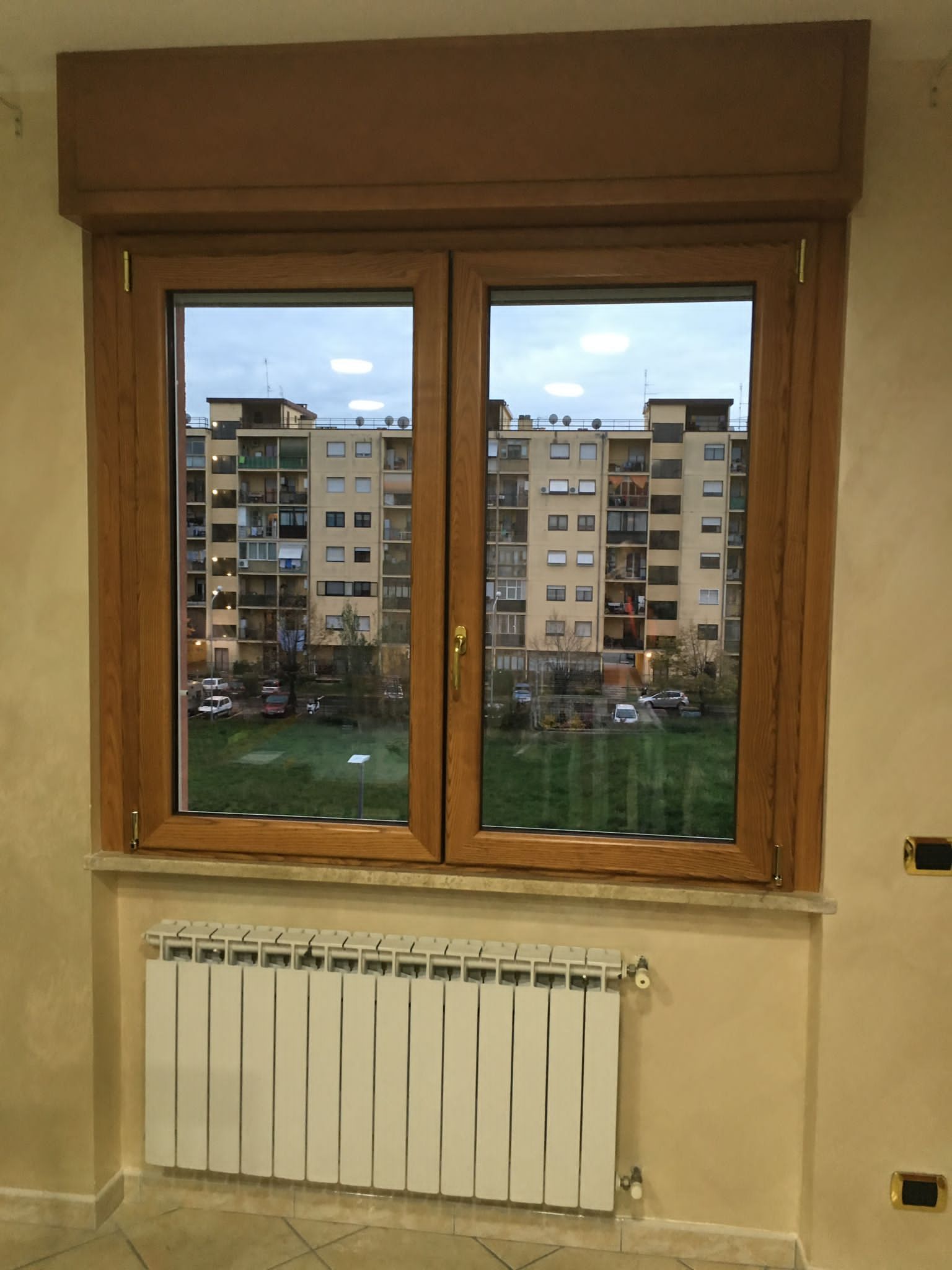 Finestra con cornice in legno sopra un termosifone bianco, affacciata su palazzi residenziali al crepuscolo.