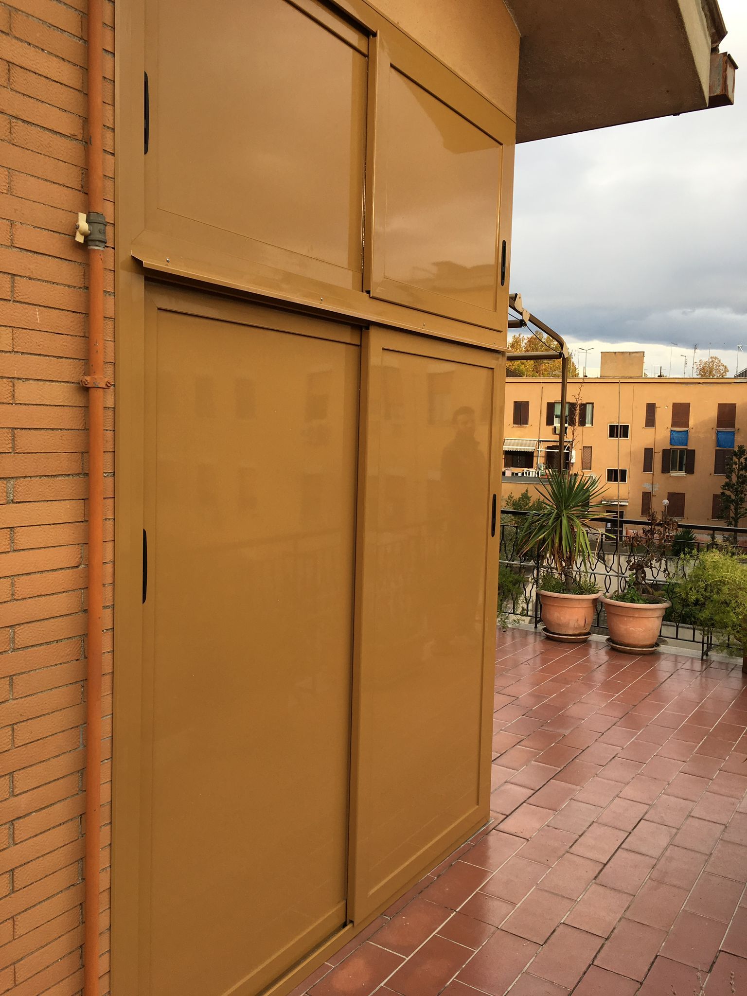 Terrazza sul tetto con persiane color beige, piante in vaso e pavimento piastrellato di rosso sotto un cielo nuvoloso.