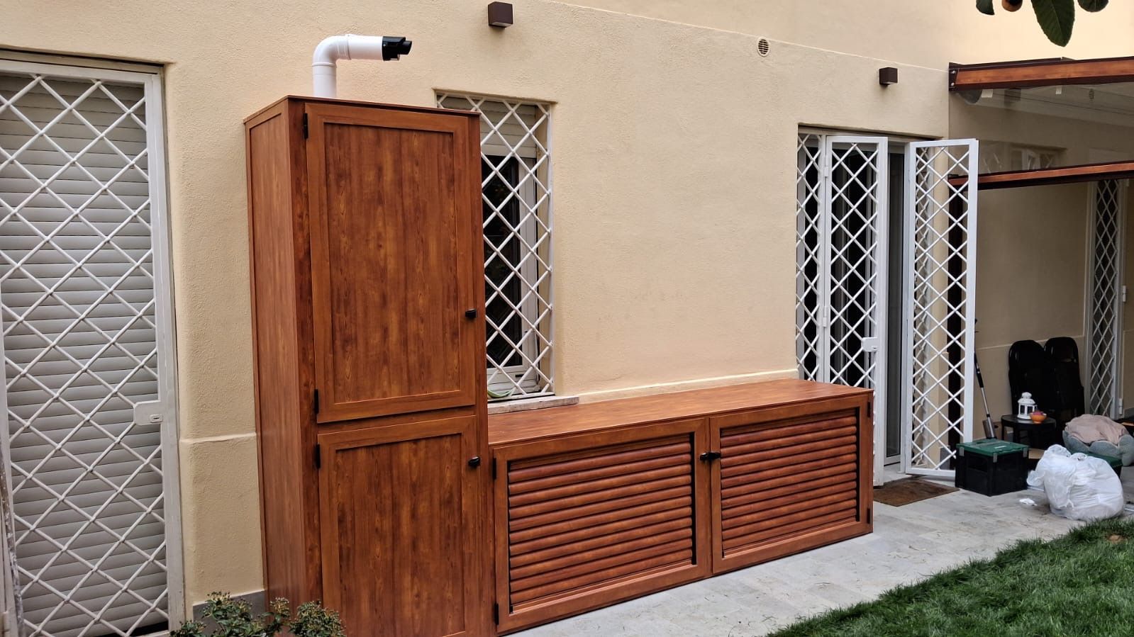 Armadietti in legno per esterni addossati a una parete beige, accanto a finestre con sbarre e una piccola porta.