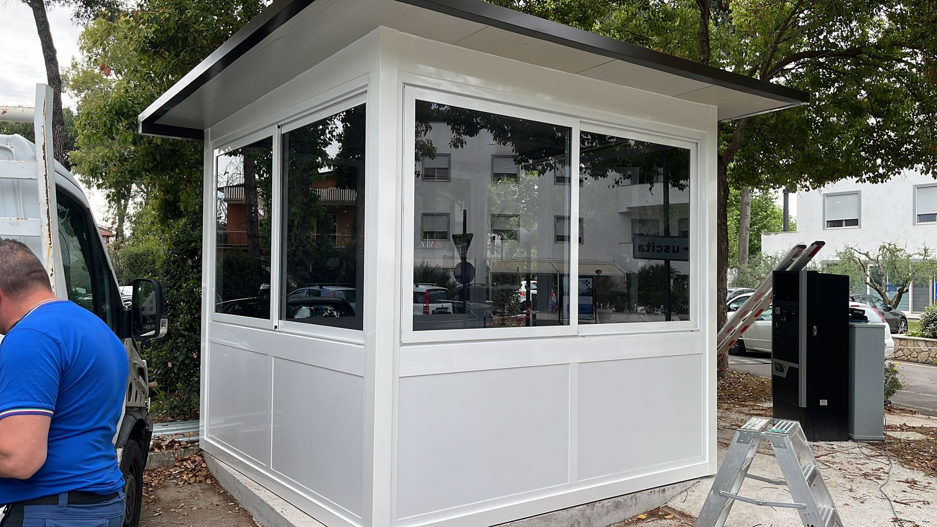 Cabina di sicurezza bianca con ampie finestre, esterna, accanto ad alberi e marciapiede.