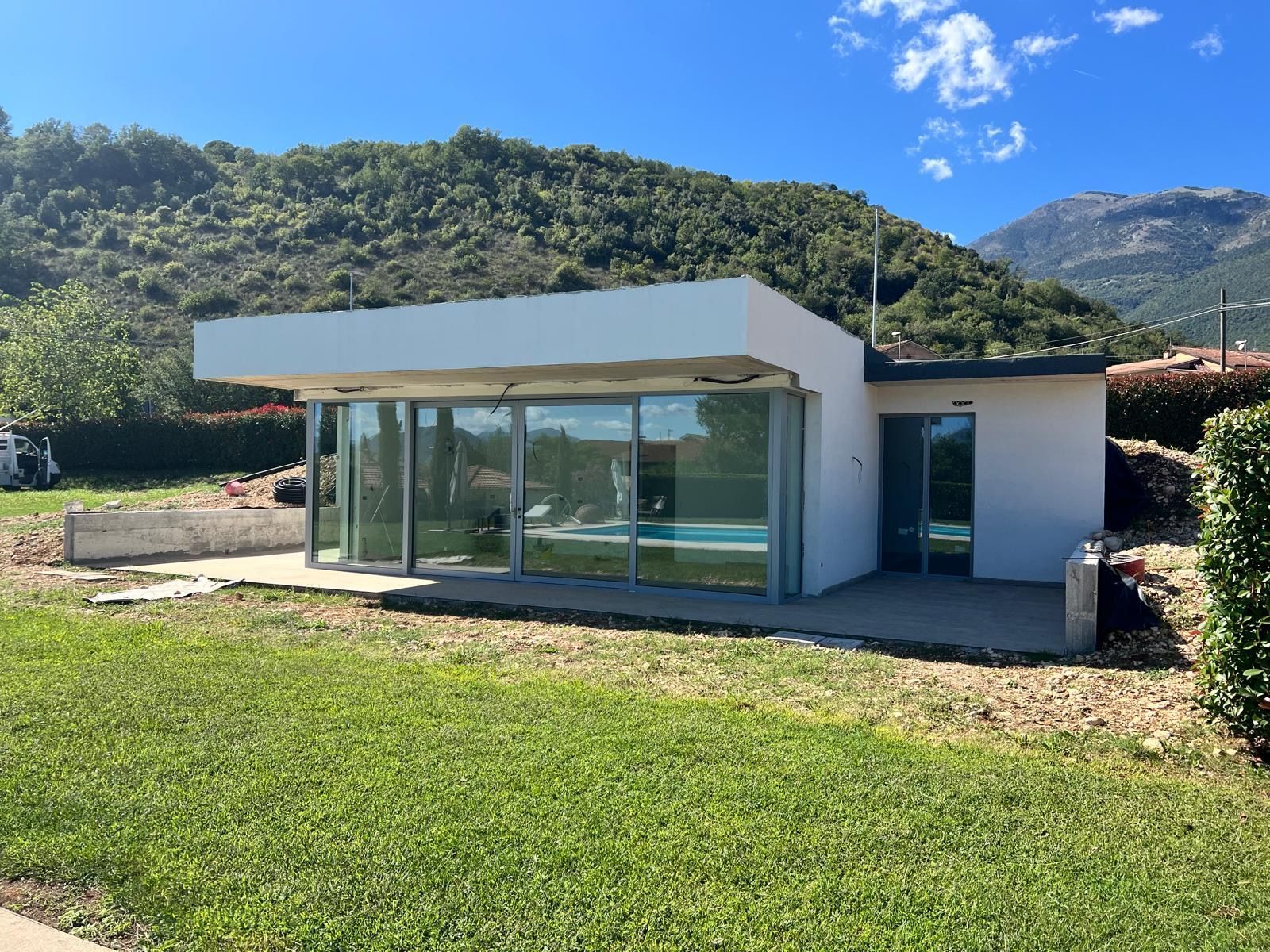 Piccola casa moderna bianca con pareti in vetro, prato verde e montagne sullo sfondo.