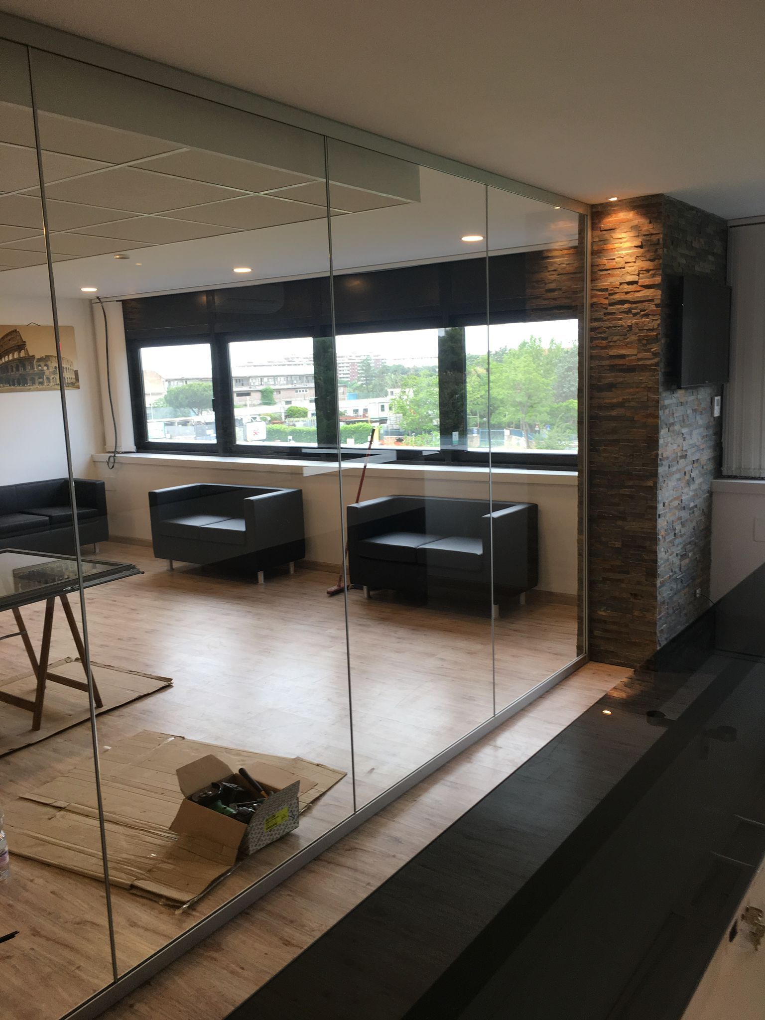 Sala relax moderna per ufficio con parete divisoria in vetro, divani neri e ampie finestre che si affacciano sul verde.
