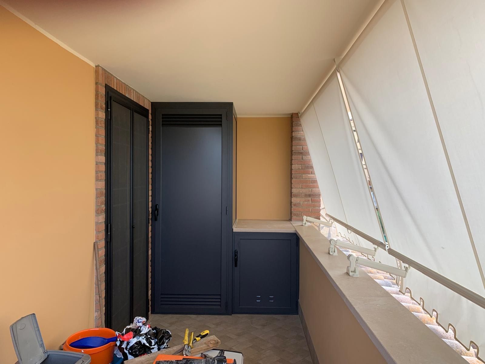 Balcone stretto con porta scura, pareti beige, tende bianche e prodotti per la pulizia sul pavimento.