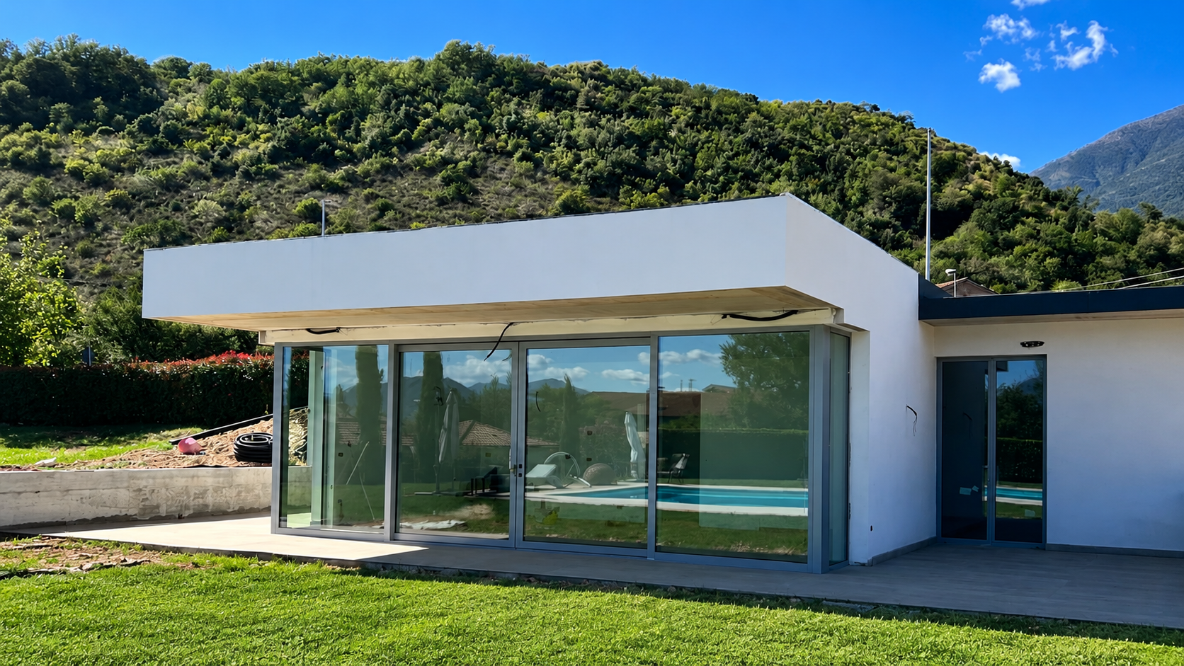 Casa moderna bianca con ampie vetrate e prato verde, incastonata tra una collina e un cielo azzurro.