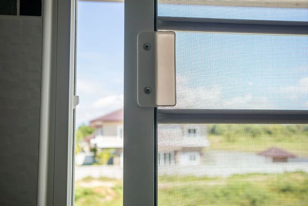 Primo piano di un fermo e del telaio di una zanzariera accanto a una vista esterna illuminata dal sole.