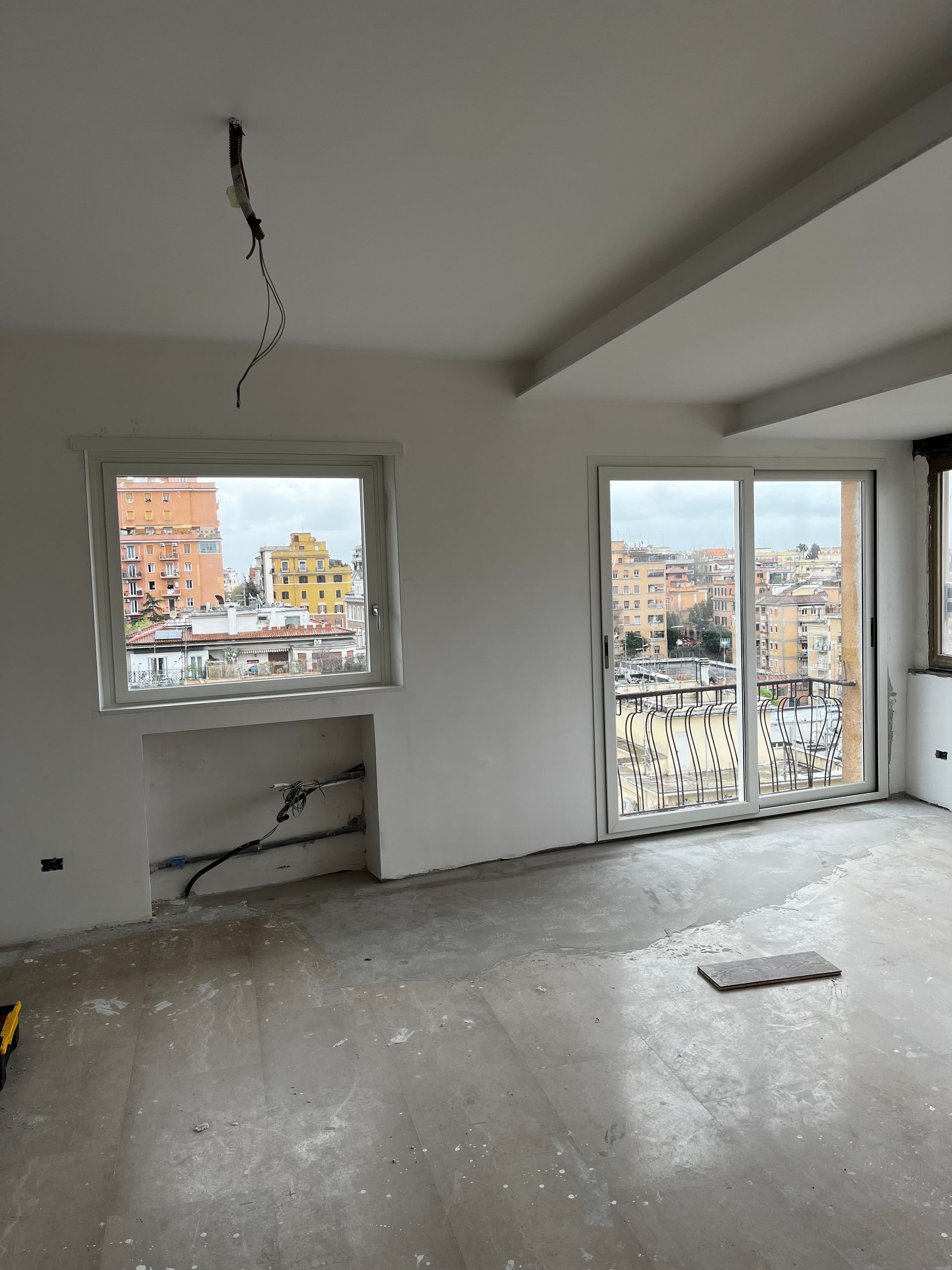 Interno di un appartamento vuoto e non rifinito, con pareti e soffitto danneggiati, pavimento in cemento e vista sulla città dalle finestre.