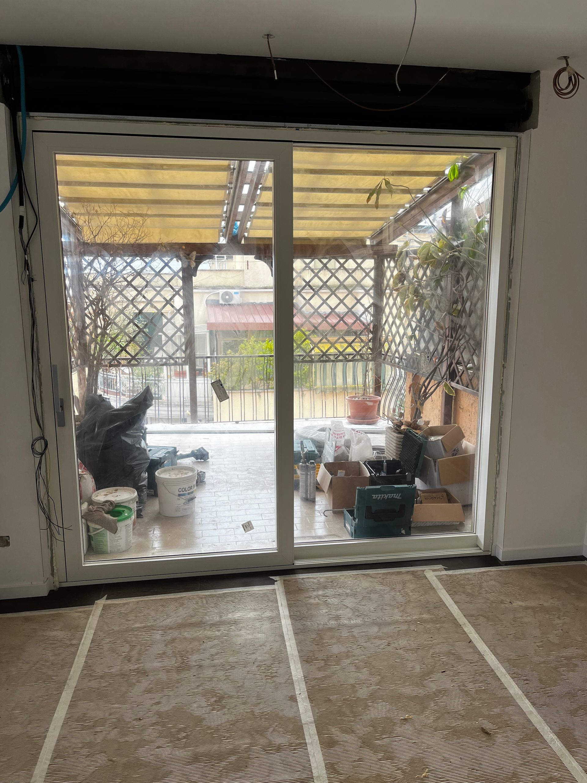 Una porta scorrevole in vetro si apre su un patio arredato con sedie e piante, visibile dall'interno attraverso un tappeto decorato.