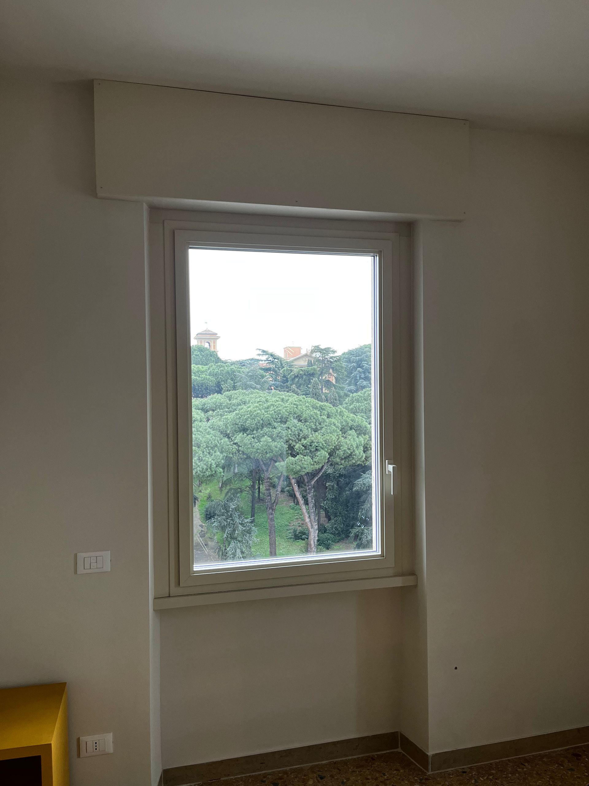Finestra con cornice bianca che si affaccia su alberi verdi in una stanza arredata in modo essenziale.