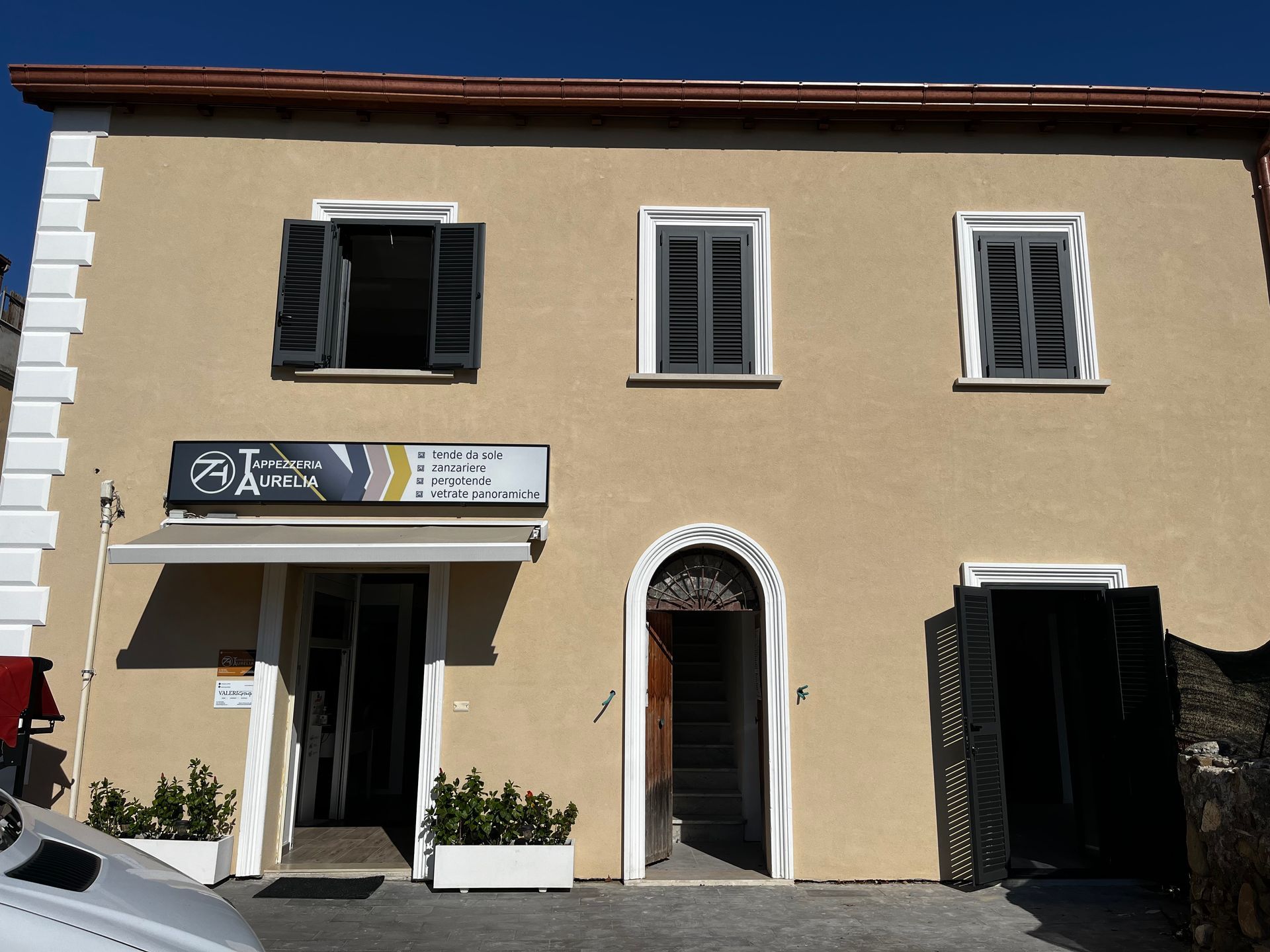 Vetrina in stucco color beige con finestre aperte, portale ad arco e persiane nere sotto un cielo luminoso