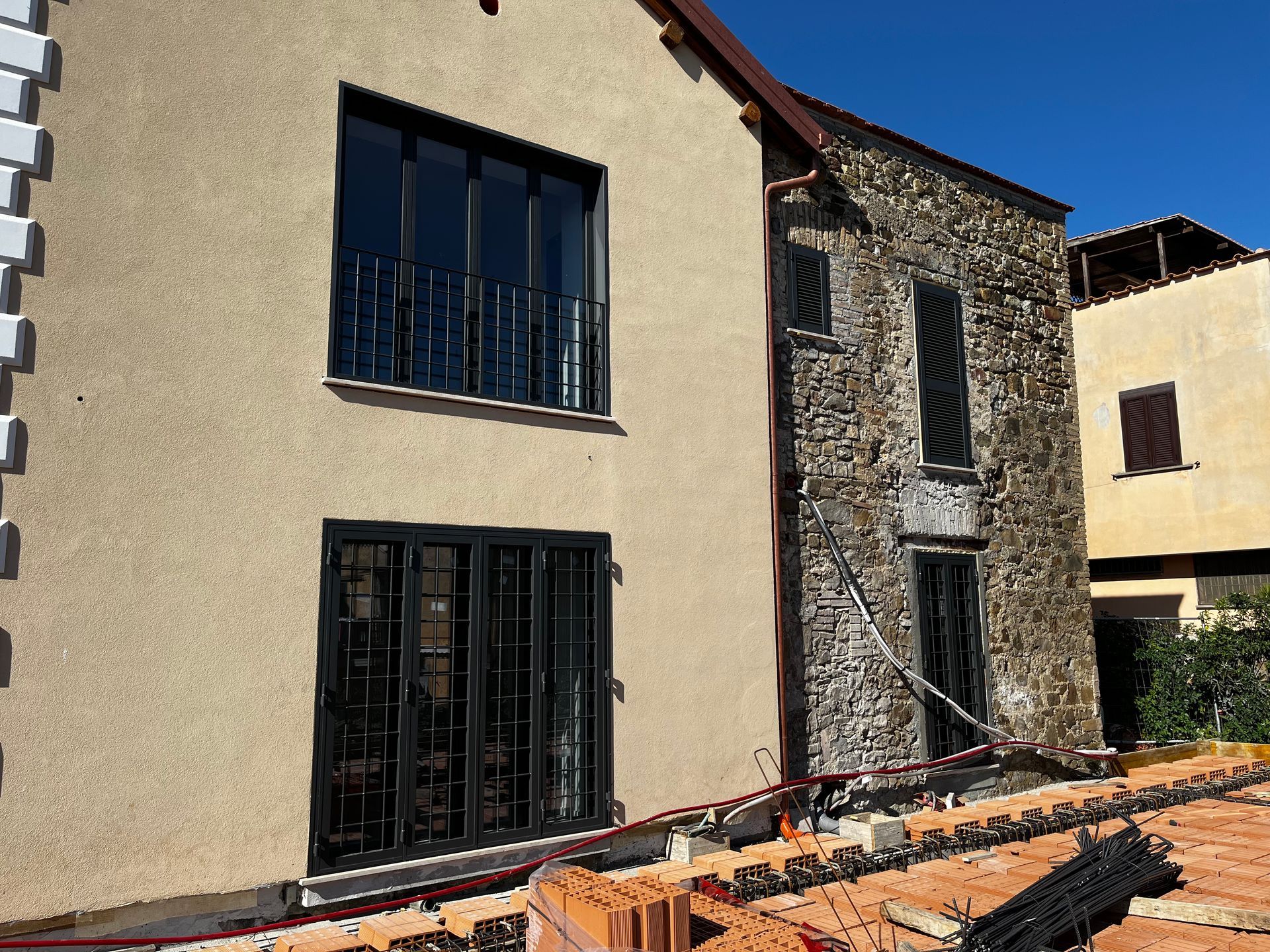 Facciata in pietra della casa con finestre con inferriate nere e tetto di tegole, accanto a un muro color pesco e aiuole.