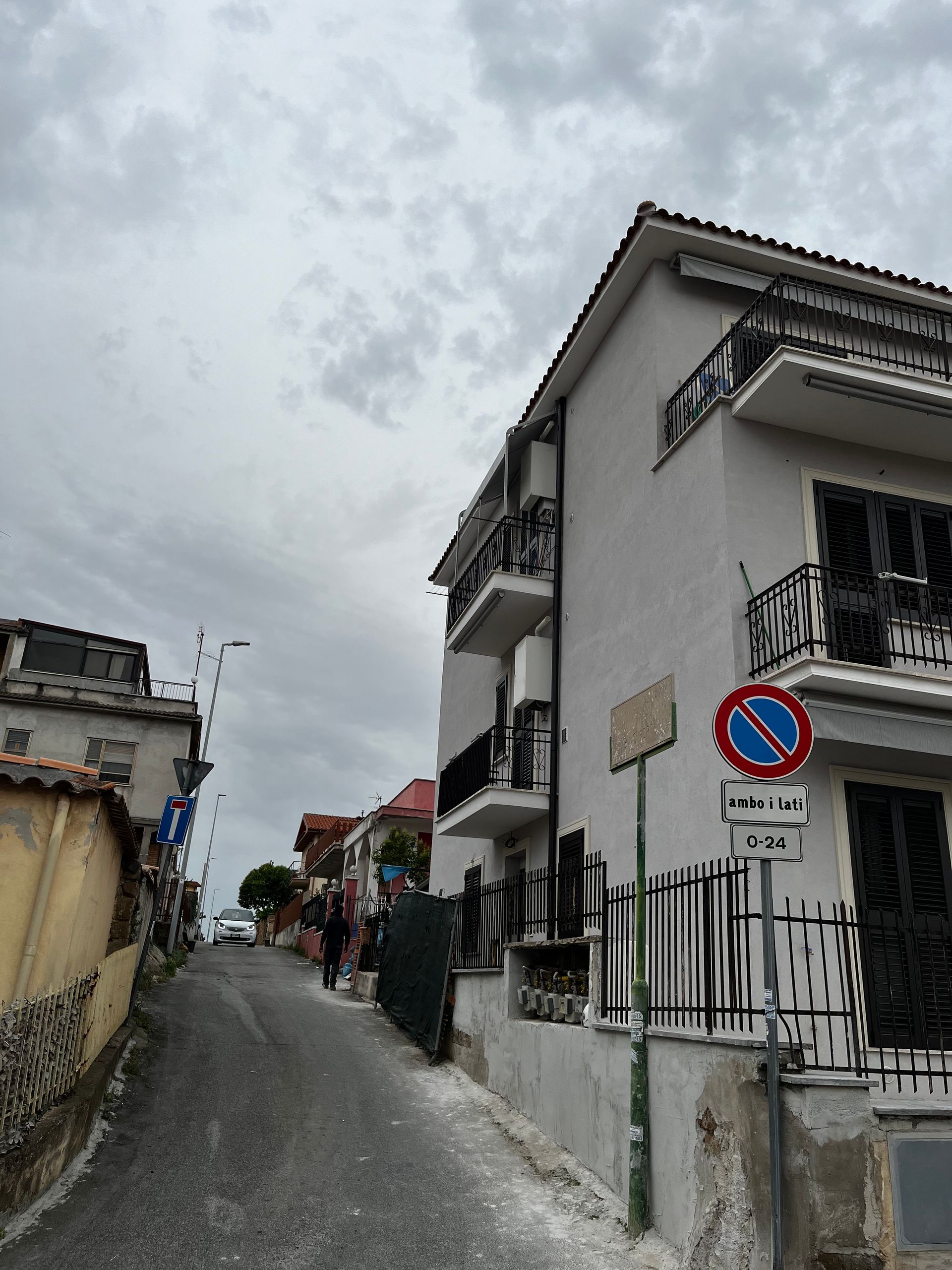 Stretta strada acciottolata tra case bianche sotto un cielo nuvoloso, con un cartello di divieto di sosta davanti.