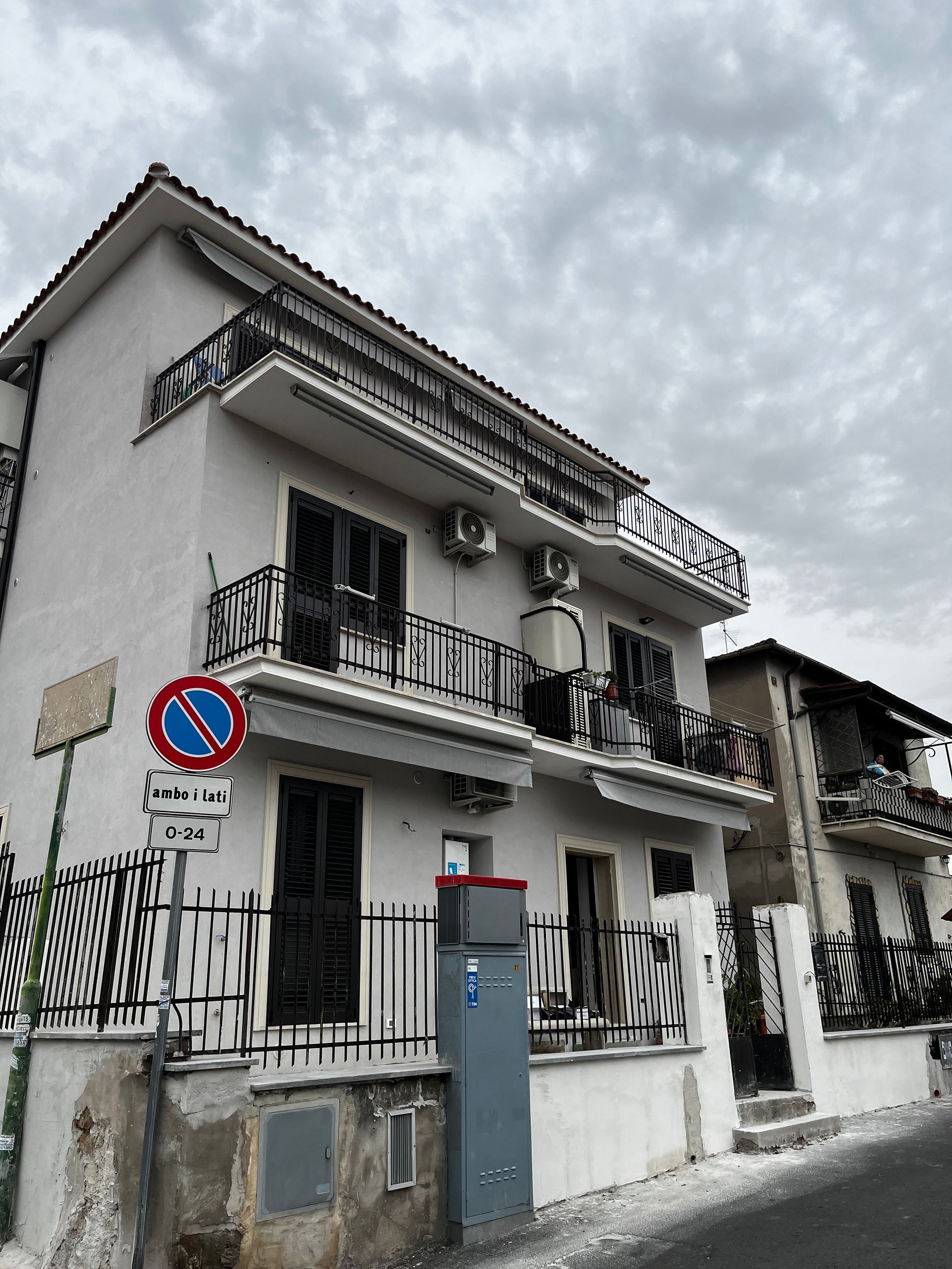 Casa bianca a due piani con balconi neri, situata all'angolo di una strada nuvolosa, con un cartello di divieto di sosta.