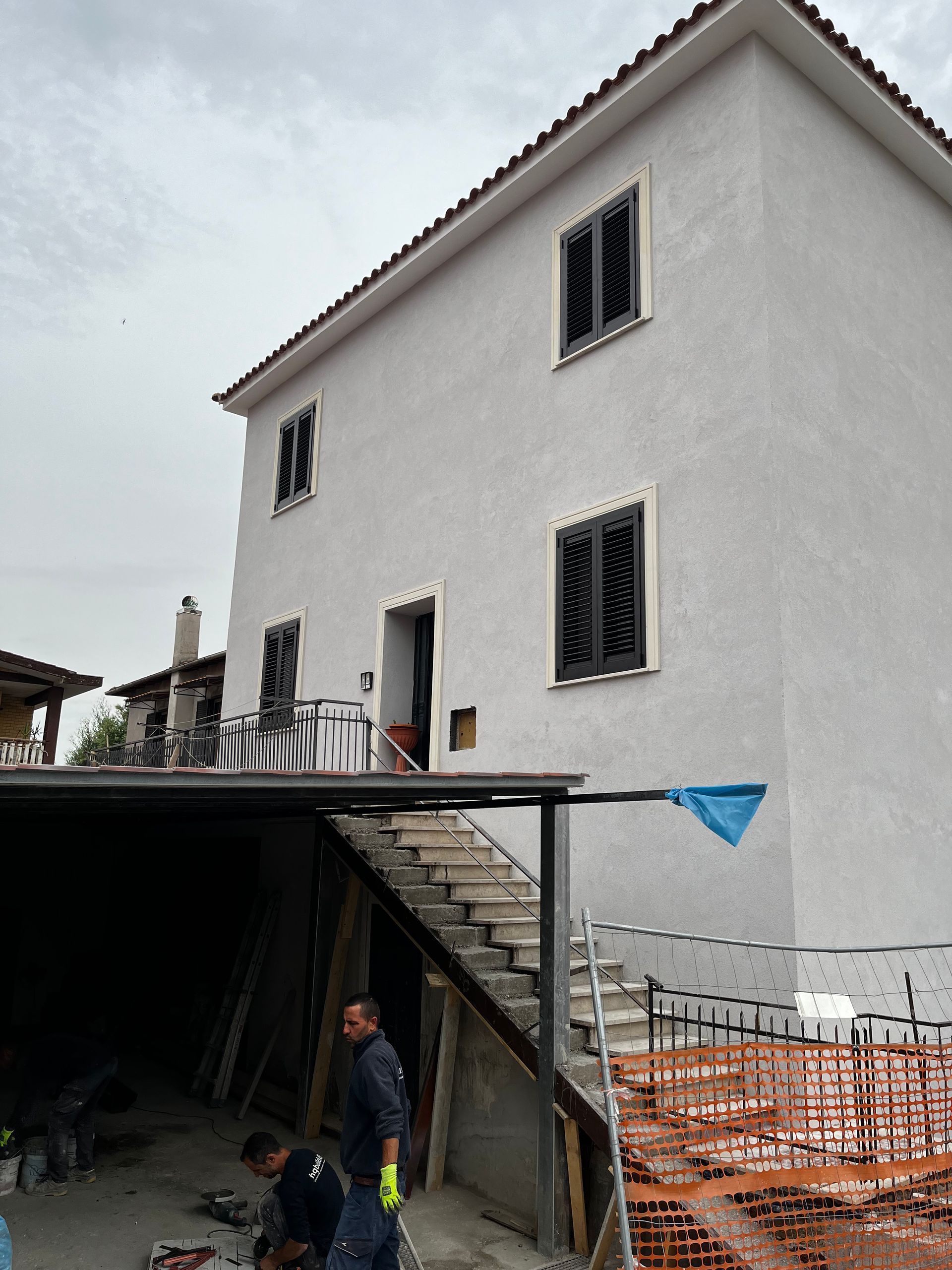 Casa bianca a due piani in costruzione con impalcature, un operaio in primo piano e un garage aperto sottostante.