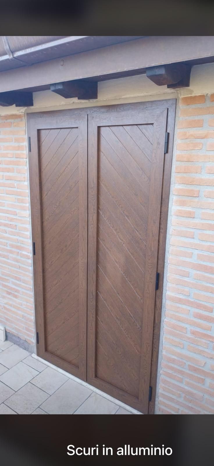 Porta doppia in alluminio marrone incassata in un muro di mattoni sotto una sporgenza del tetto.