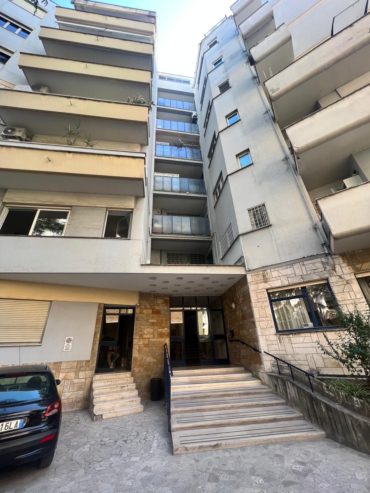 Ingresso di un condominio con scale, balconi e auto parcheggiata di fronte.