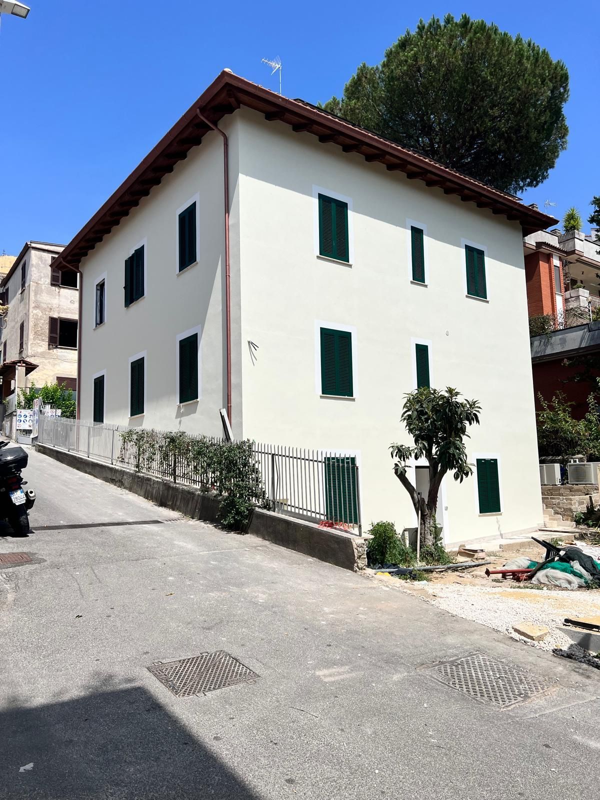 Casa bianca ad angolo su una strada in pendenza, con persiane verdi e un grande albero alle spalle.