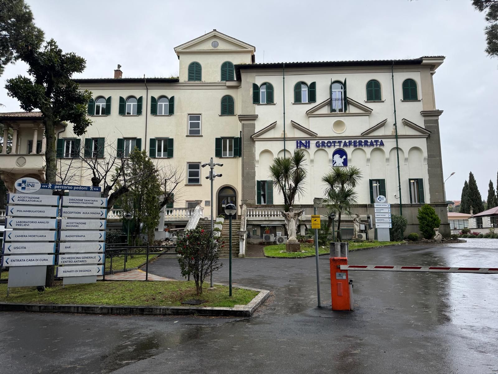 Vista sotto la pioggia di un edificio bianco con persiane verdi e un cartello d'ingresso al vialetto.