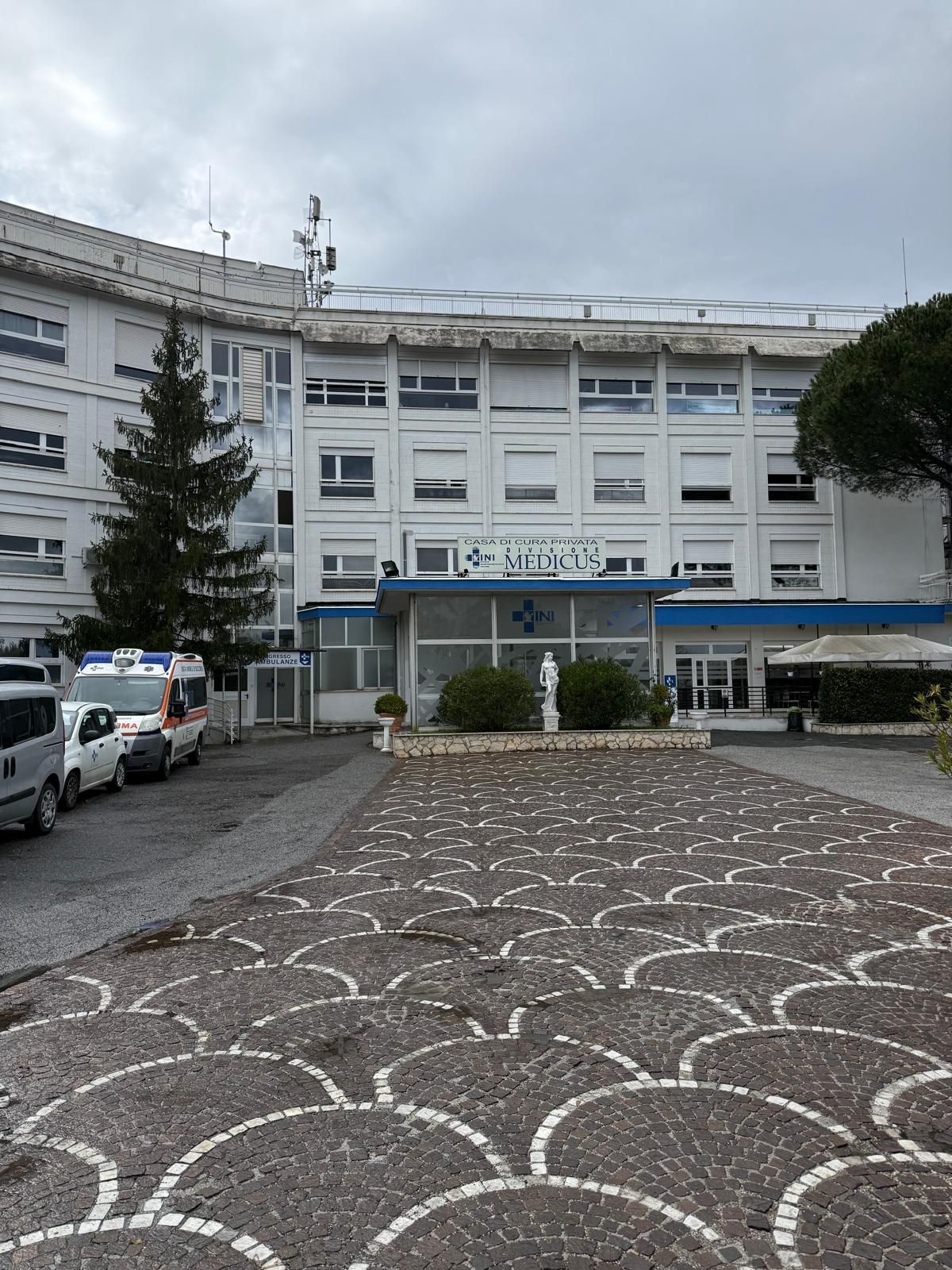 Grande edificio ospedaliero bianco con auto parcheggiate e un cortile in pietra decorato sotto un cielo nuvoloso