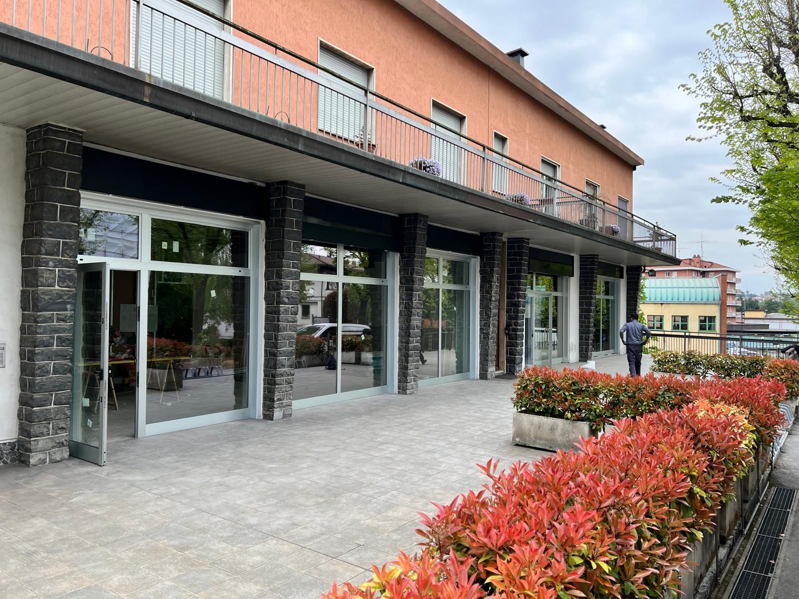 Ingresso moderno dell'edificio con porte a vetri, colonne in pietra e aiuole di fiori rossi lungo un vialetto lastricato.
