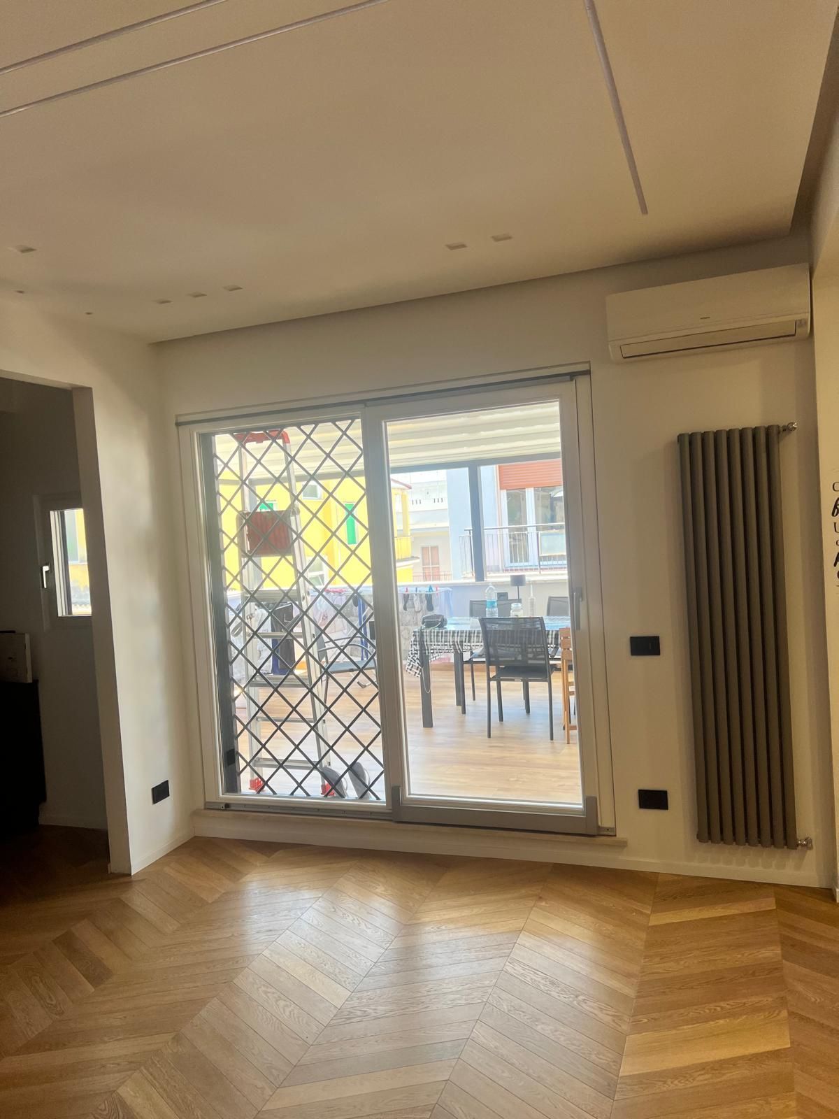 Stanza vuota con pavimento in legno a spina di pesce, ampia porta a vetri e balcone con vista sull'esterno.
