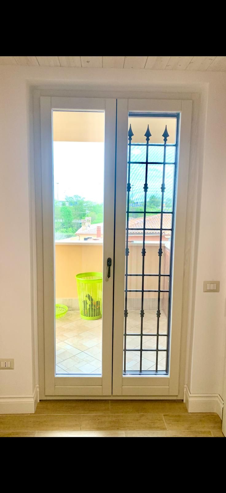 Porte finestre bianche con vetro satinato che si aprono su un balcone con piante e un cancello di sicurezza in metallo.