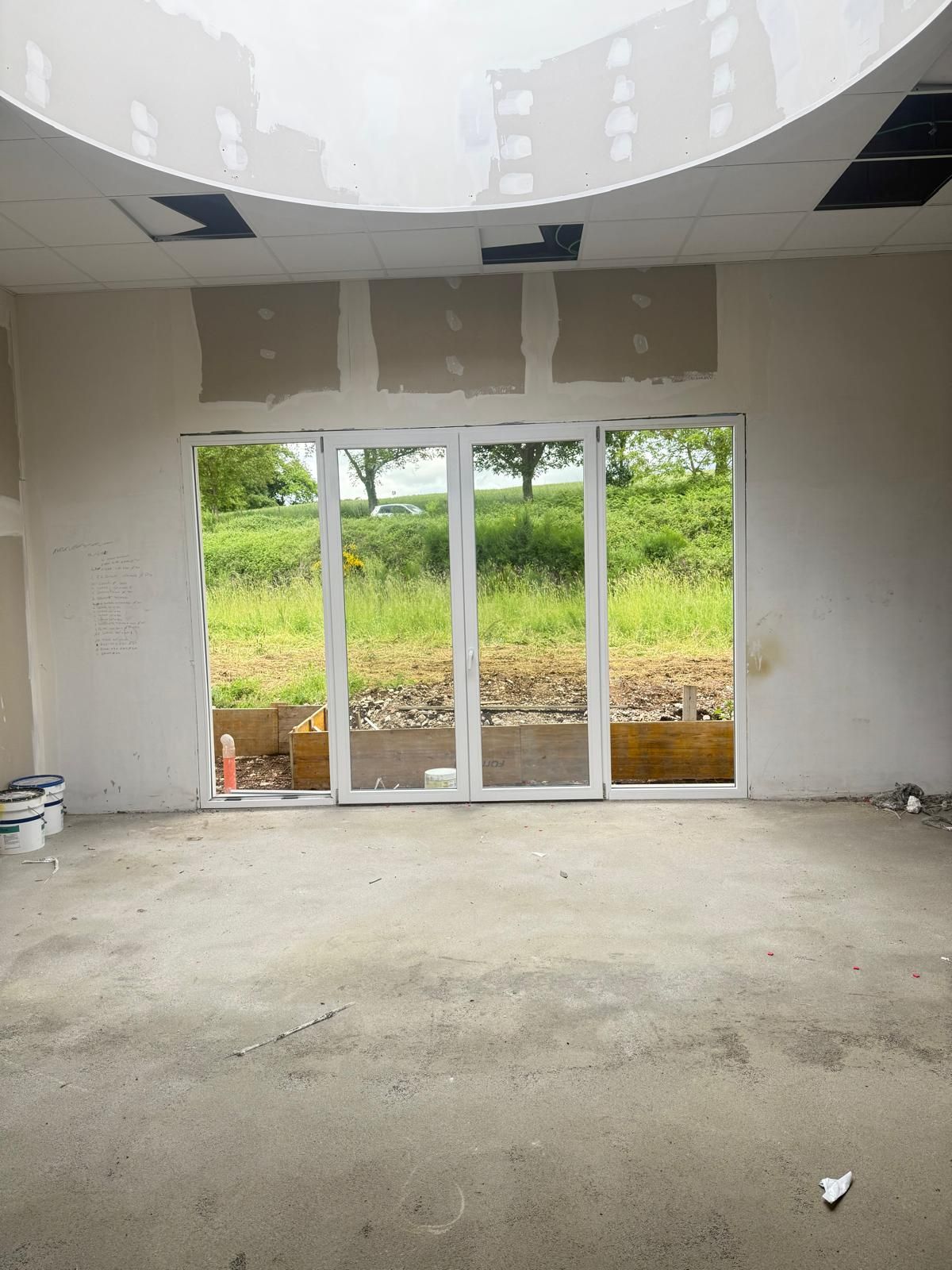 Stanza non rifinita con porte a vetri che si aprono su una vista esterna verde e soffitto curvo in costruzione