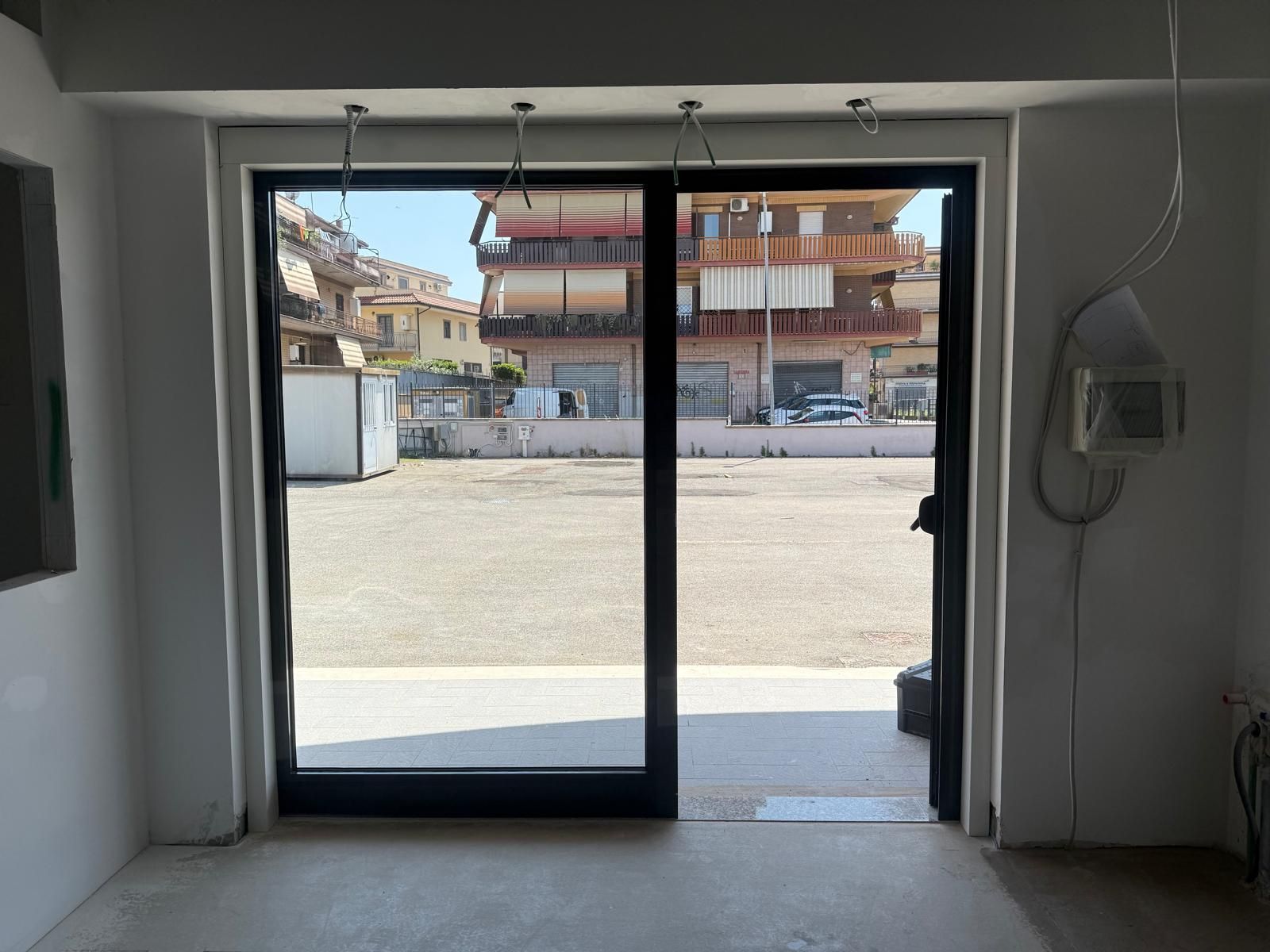 Vista attraverso porte a vetri su una strada soleggiata e sugli edifici, con un interno grigio in primo piano.