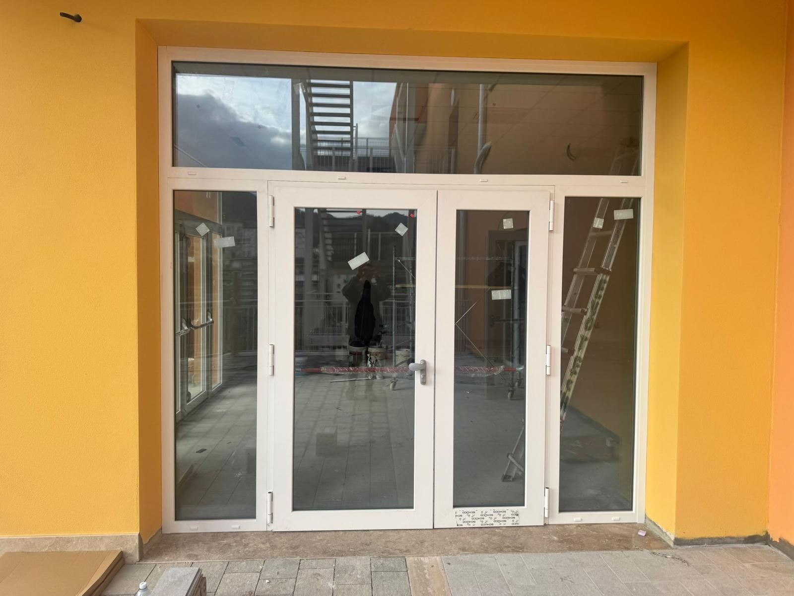 Ingresso dell'edificio giallo con doppie porte a vetri bianchi e finestre a sopraluce, attualmente in costruzione.