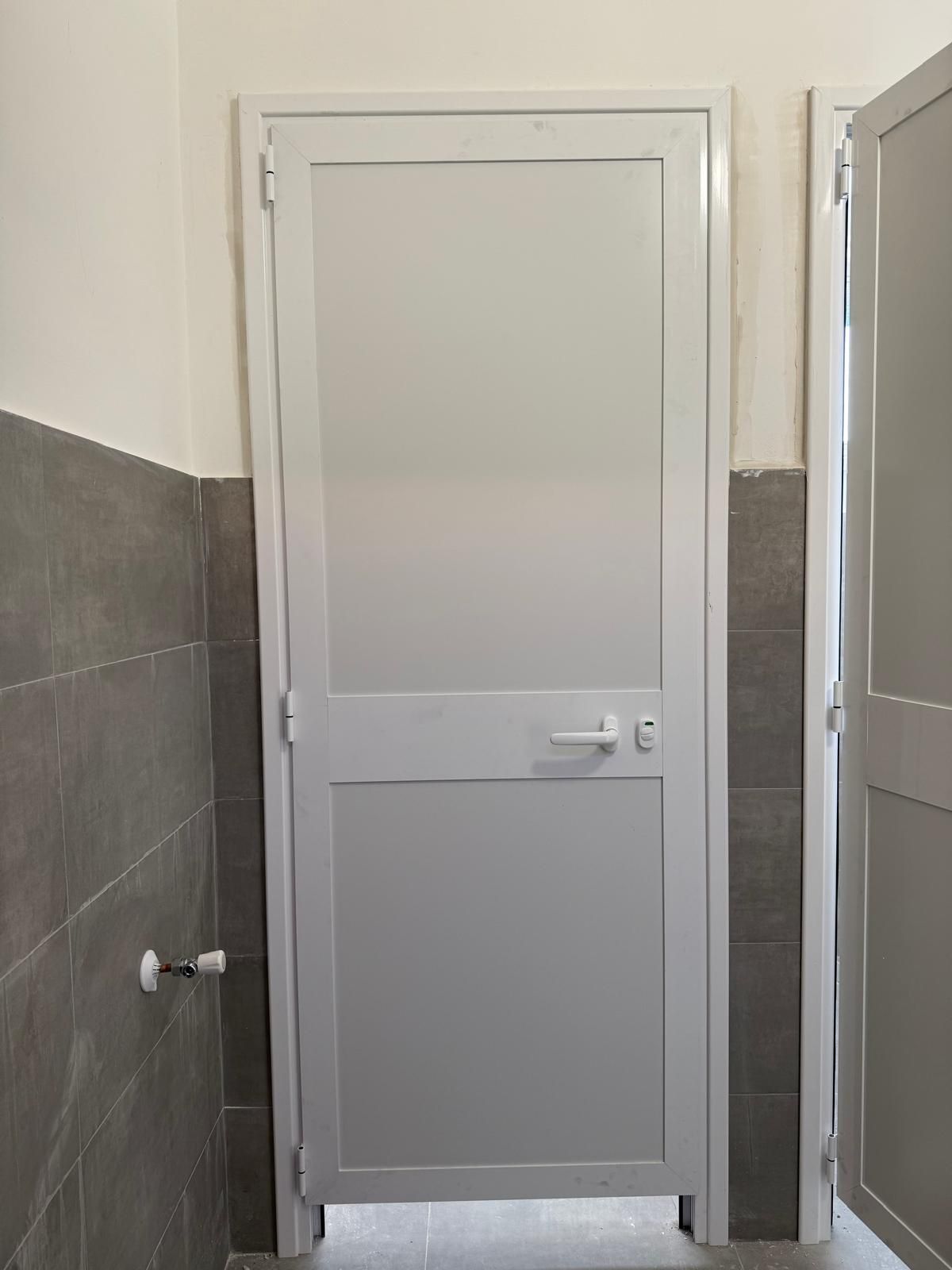Porta bianca con un piccolo pannello inferiore in un corridoio interno piastrellato di grigio.