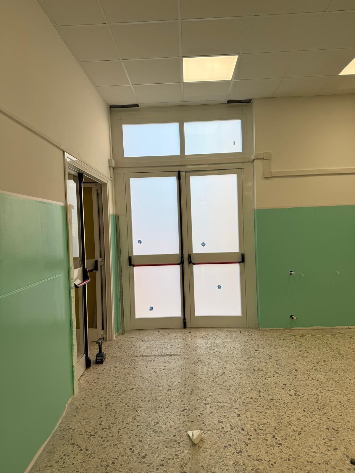 Corridoio vuoto con pareti inferiori verdi, pavimento in terrazzo e doppie porte a vetri in fondo.